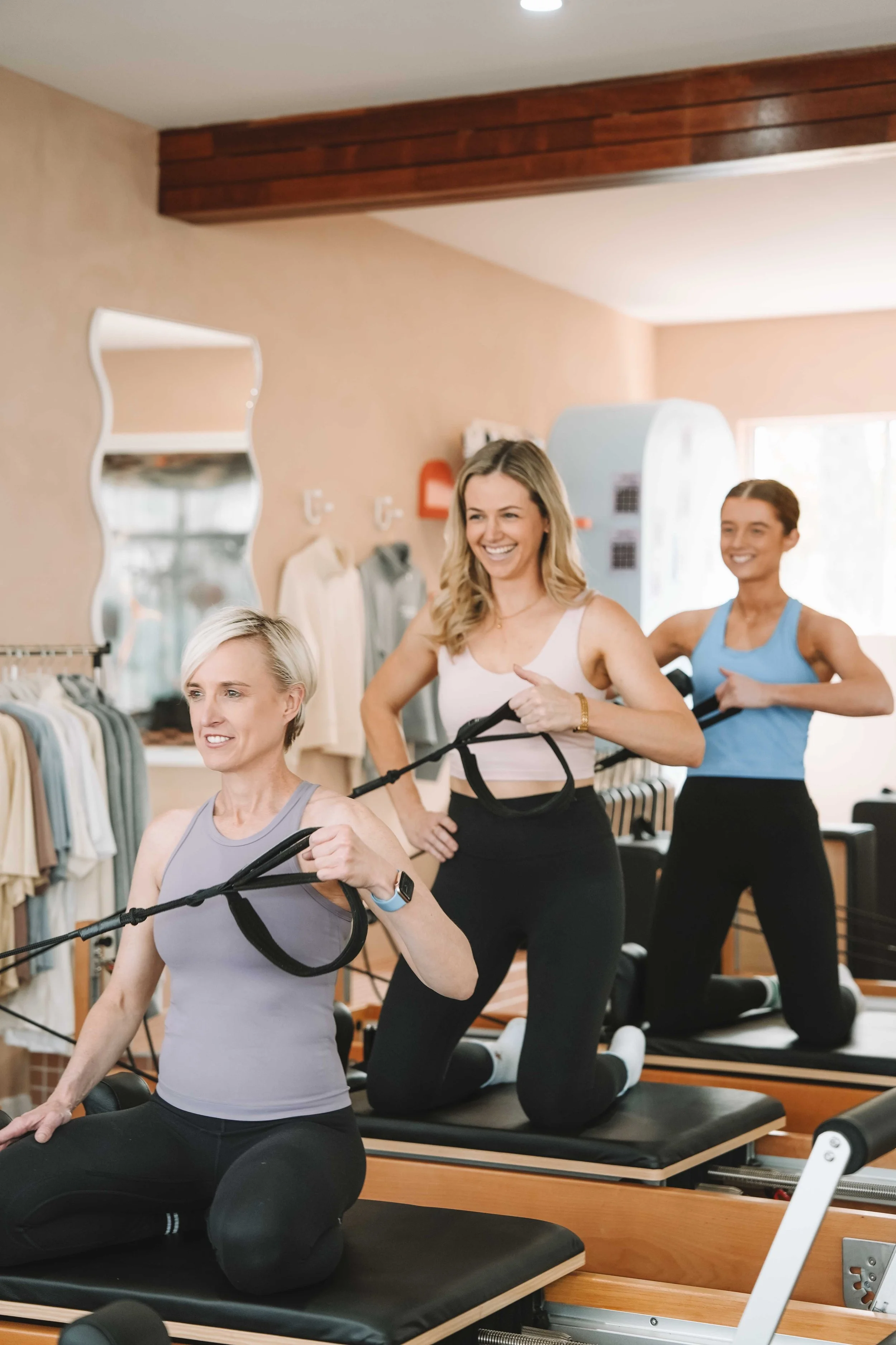 Three women exercising with resistance bands on reformer machines in a fitness studio, smiling and appearing happy.