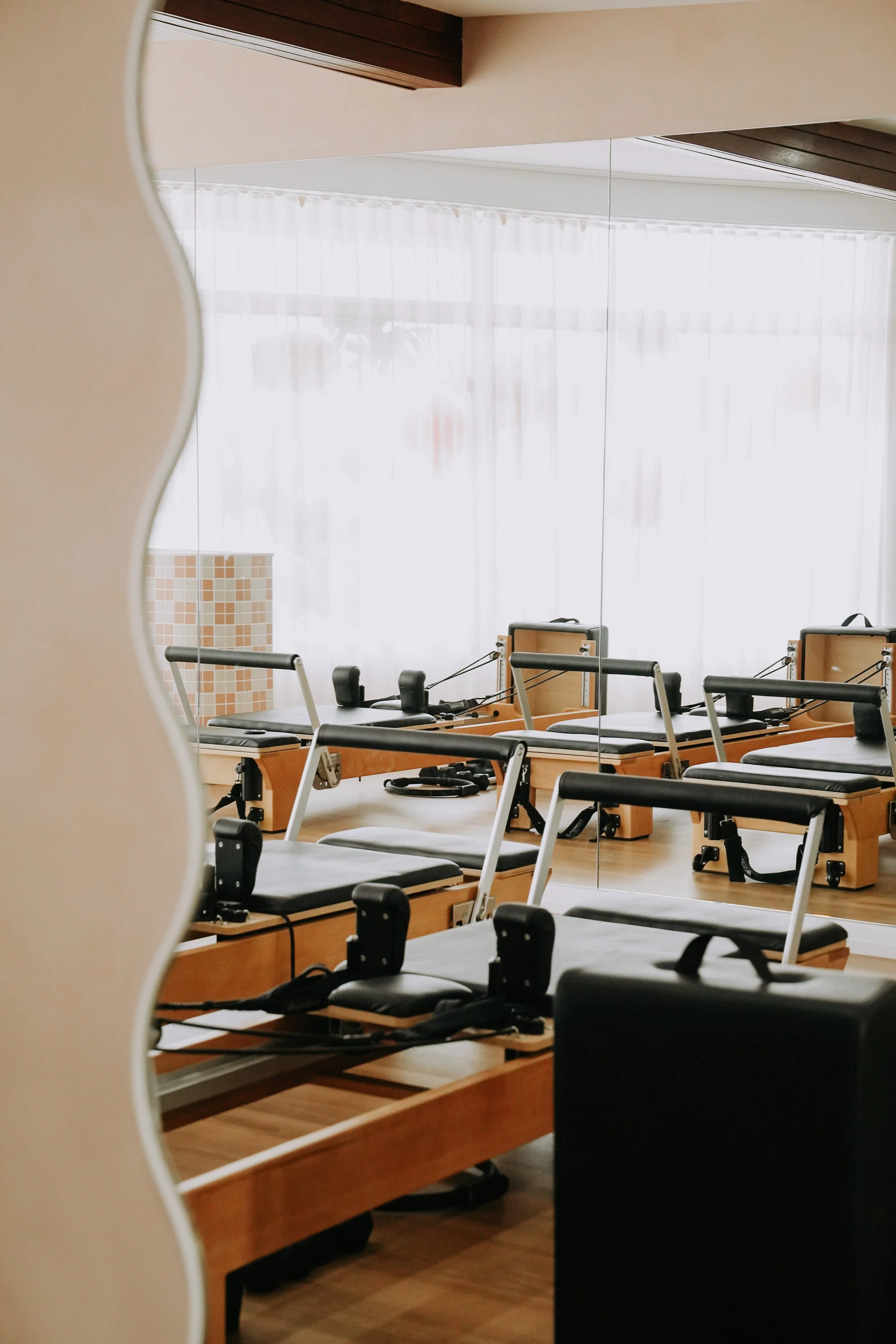 Pilates reformer machines reflected in a large mirror, with a sheer curtain and natural light in the background.