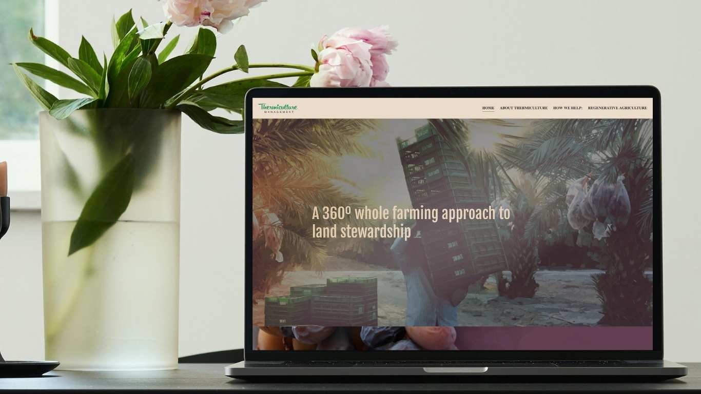 Laptop screen displaying a website about regenerative agriculture with a background of palm trees and sunlight. A vase with pink flowers and green leaves is on the left side of the laptop.