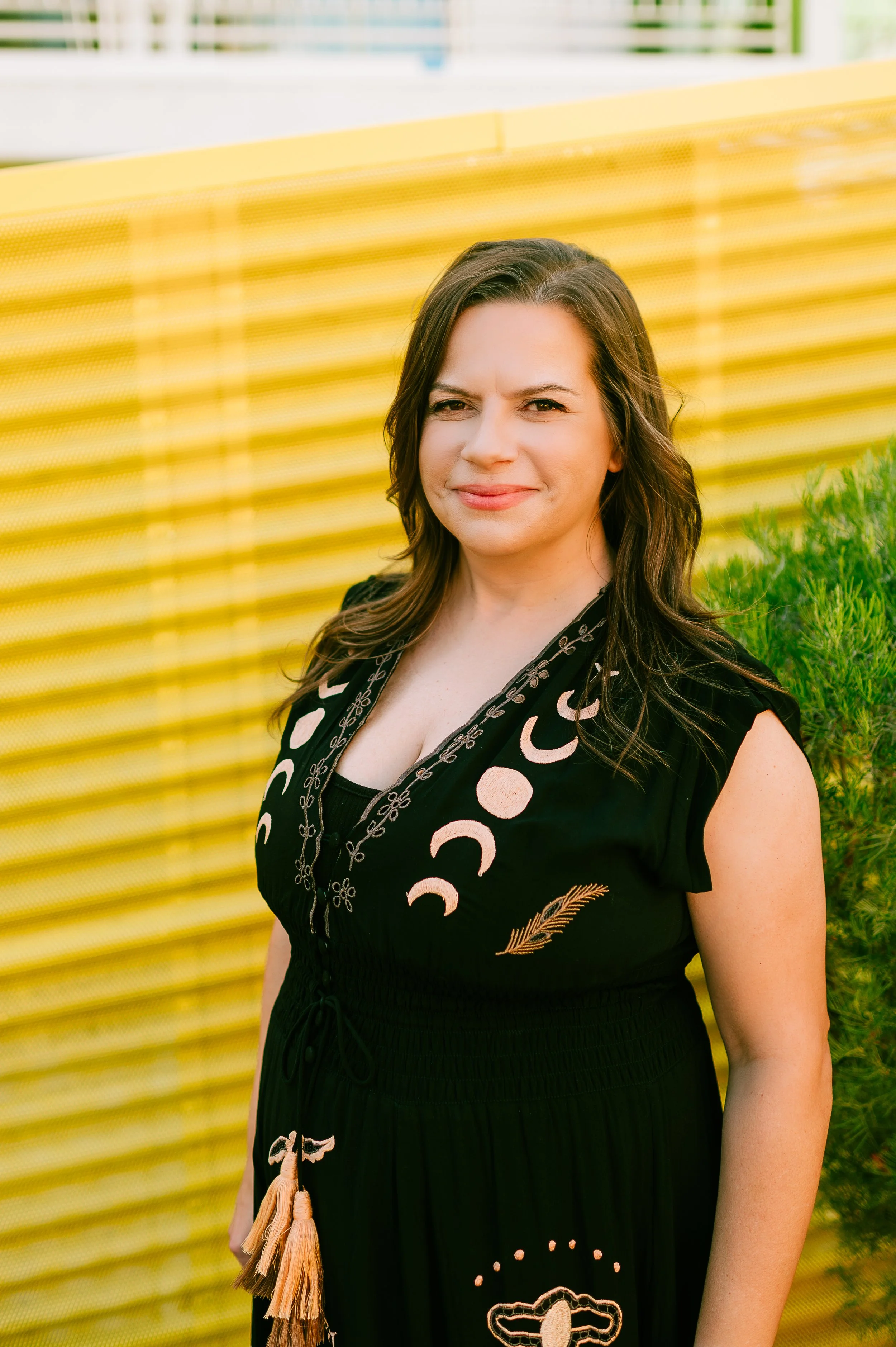A woman with brown hair wearing a black dress with moon and feather patterns, standing outdoors near a green bush and a yellow wall.