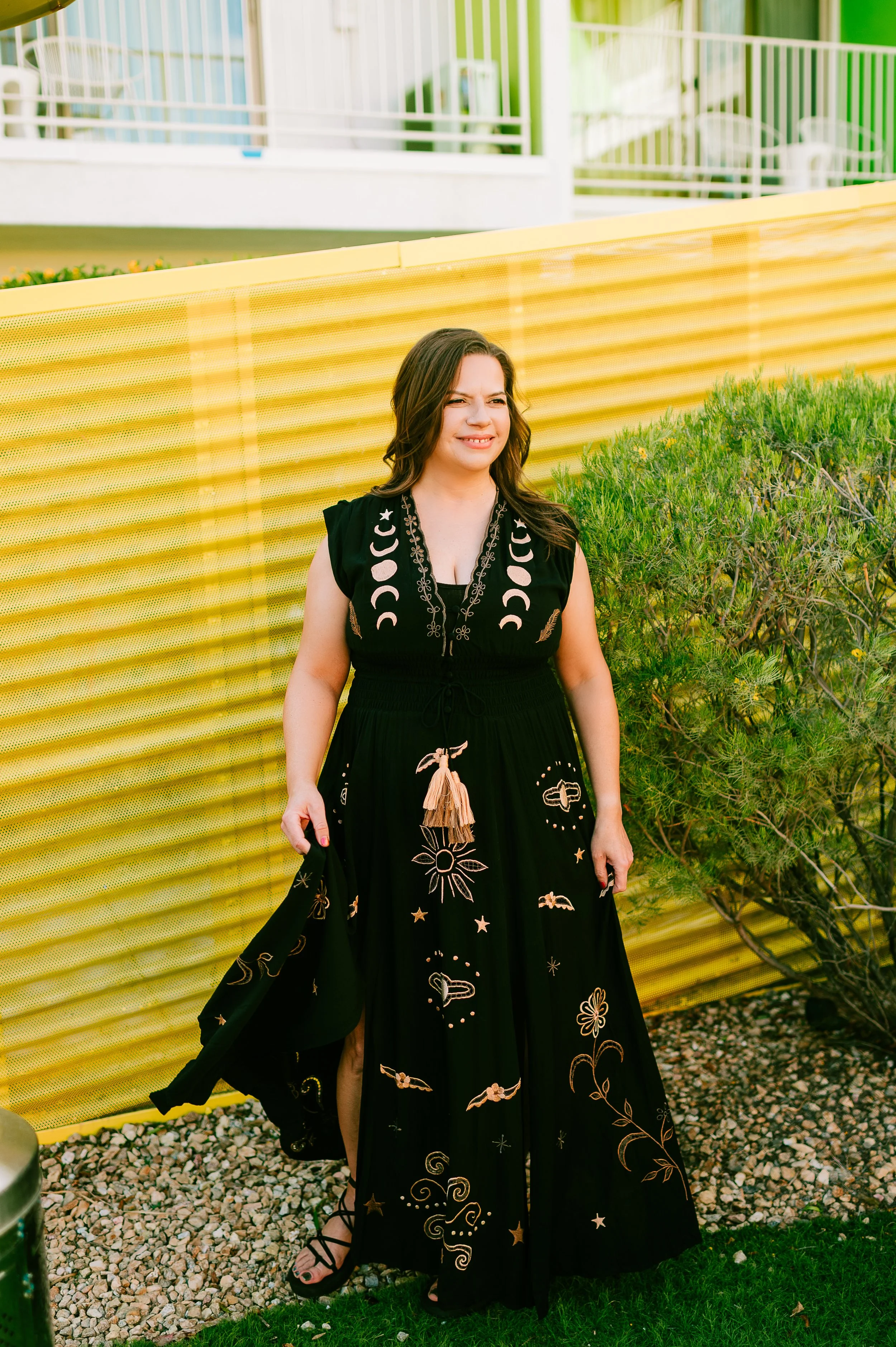 Woman in a black dress with celestial and floral embroidery, standing outdoors near a yellow fence and green bush, making a playful face.
