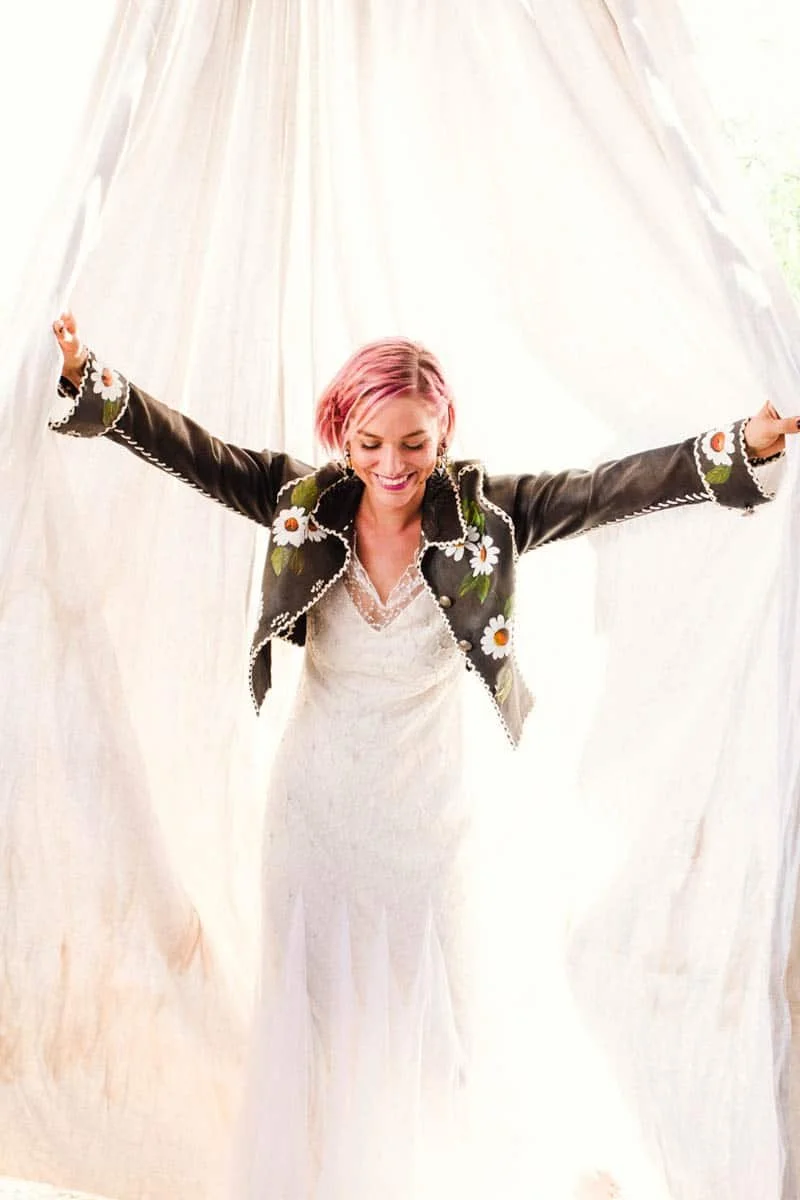 A woman with pink hair smiling and spreading her arms wide, standing in front of a white curtain or sheet.