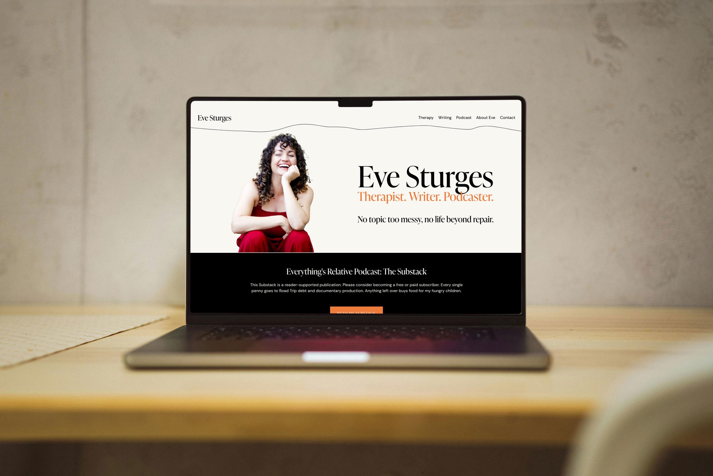 Laptop on a wooden desk displaying a website with a photo of a woman in red sitting and smiling, with text introducing her as Eve Sturges, a therapist, writer, and podcaster.