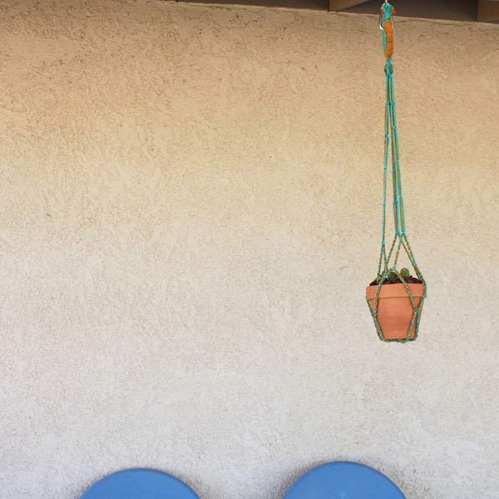 A hanging plant pot in a decorative macrame hanger on a wall, with two blue chairs at the bottom of the image.