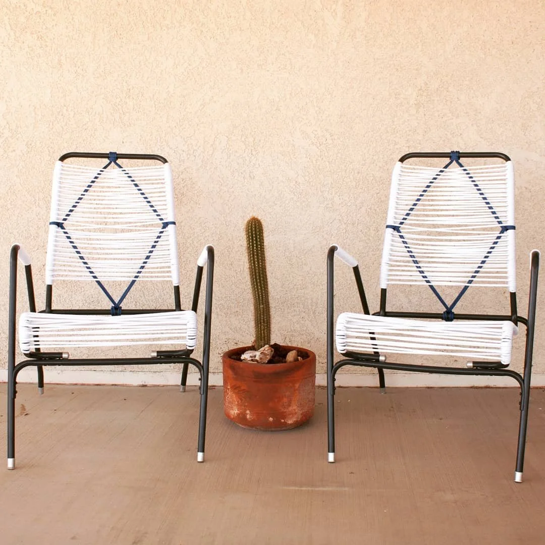 Two white outdoor chairs with black frames and striped white and blue cushions on a beige concrete patio, flanking a potted cactus in a terracotta pot against a beige textured wall.