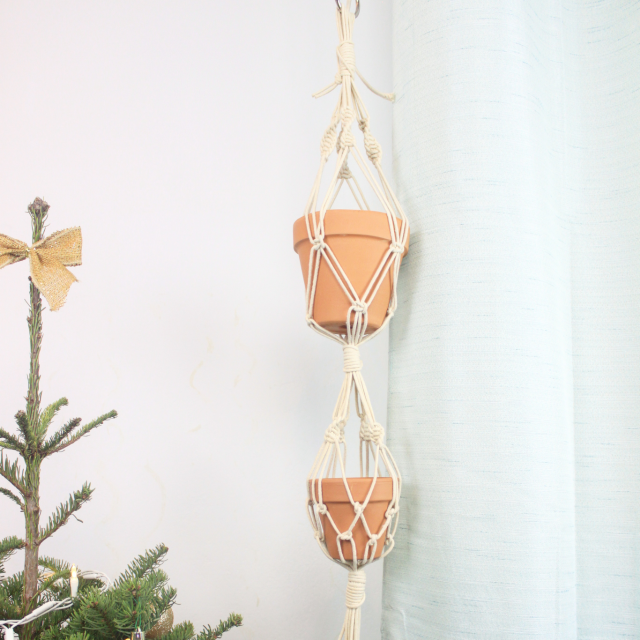 Two terracotta plant pots hanging in a macrame plant hanger against a plain light-colored wall and near a light-colored curtain, with part of a decorated Christmas tree visible in the lower left corner.