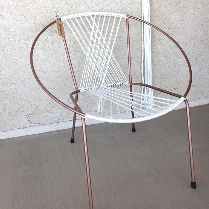 Modern chair with a circular copper-colored metal frame, white macrame seat and backrest, placed on a concrete floor against a textured wall.