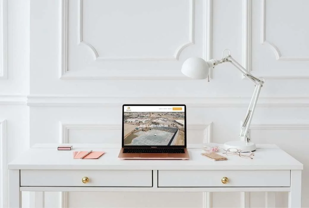 White desk with a laptop, pink notebooks, glasses, a lamp, and a phone in a bright, minimalistic room.