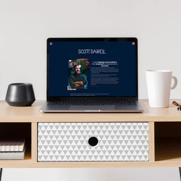 Laptop on a wooden desk displaying Scott Darrell's profile, with a black container, white mug, and a pen nearby.