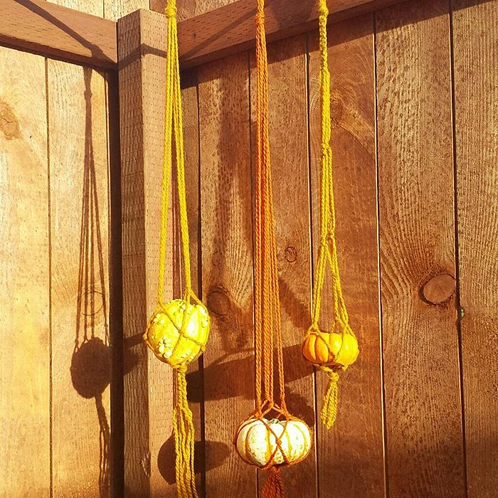 Four decorative gourds in yellow and orange are hanging with macrame-style knotted strings against a wooden wall.