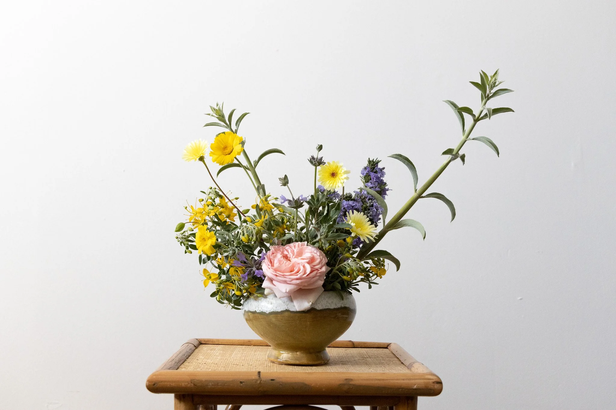 A ceramic vase containing a colorful arrangement of various flowers, including yellow, purple, and pink blossoms, placed on a wooden table against a plain white background.