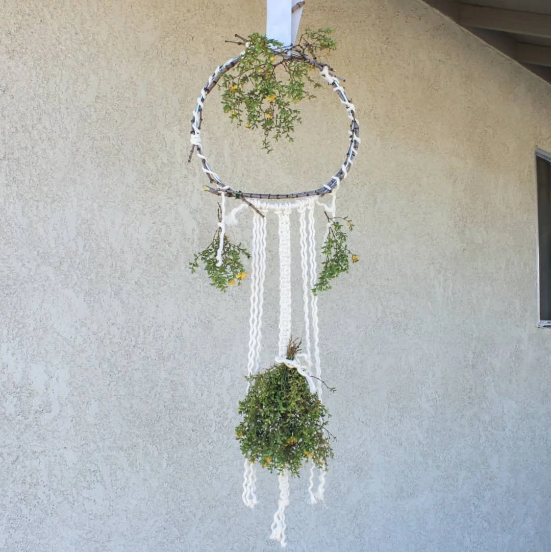 A hanging macrame design made with a metal hoop wrapped in white rope, hanging in front of a wall. It has small green creosote plant branches with yellow flowers hanging from it.