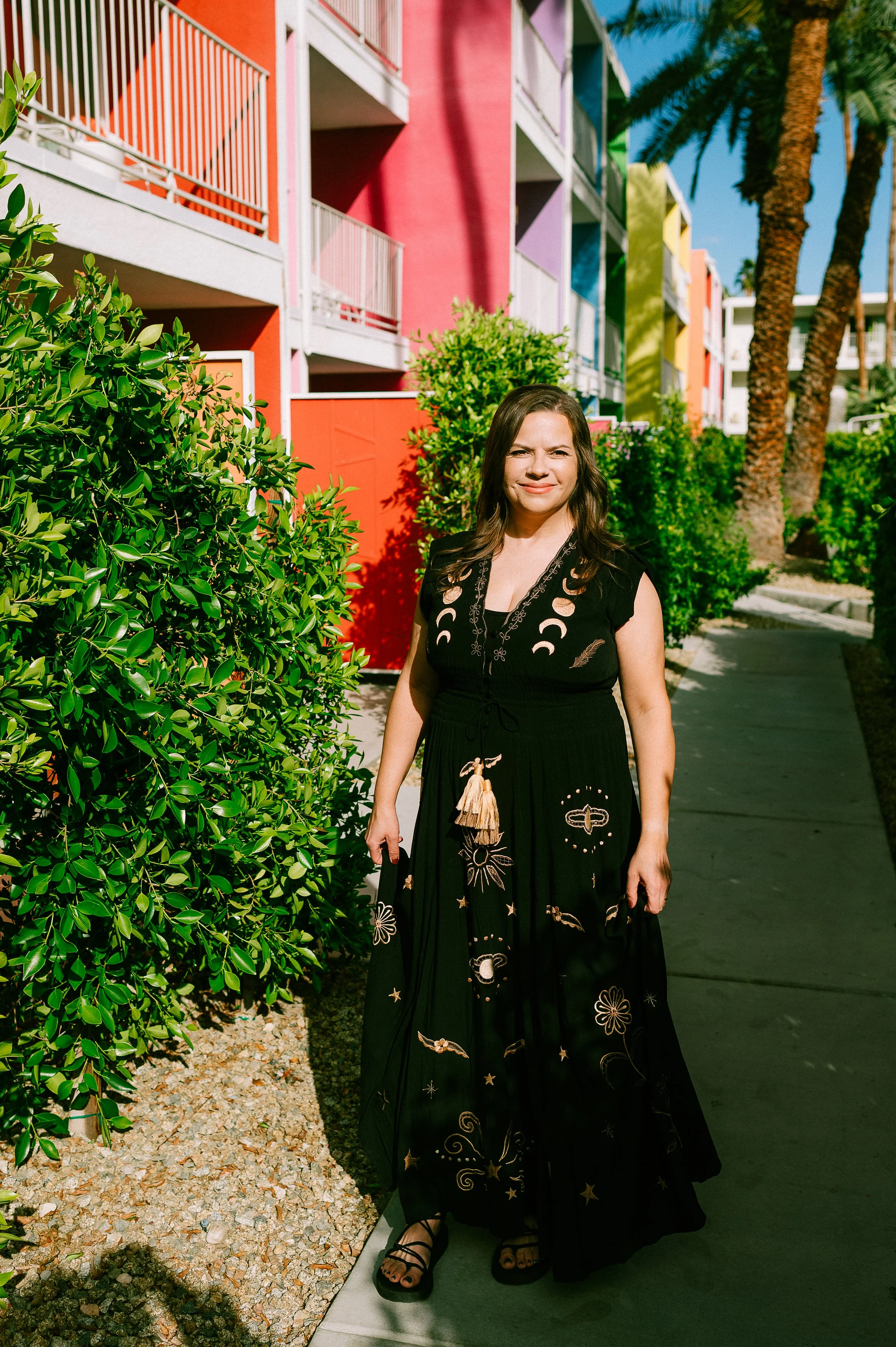 A woman in a black dress with moon and star patterns standing on a sidewalk next to lush green bushes, with colorful apartment buildings and palm trees in the background under a clear blue sky.