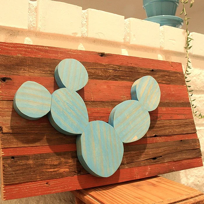 Decorative piece made of stacked circular wood pieces painted light blue with white stripes representing cactus pads, arranged on a rustic wooden plank leaning against a wall.
