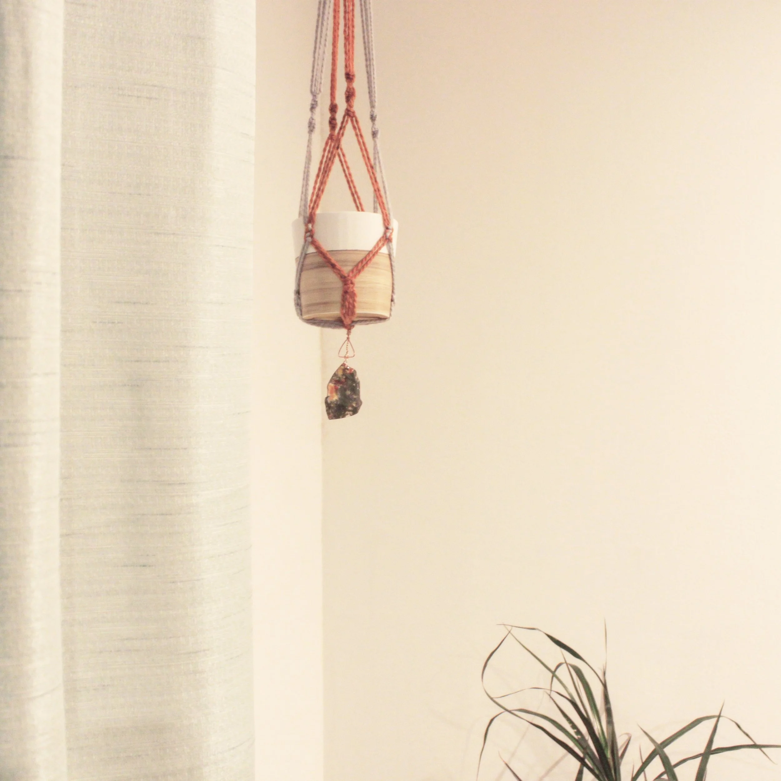 A hanging planter with a pot and a decorative stone, suspended by braided cords, near a beige wall and a plant in the bottom right corner.