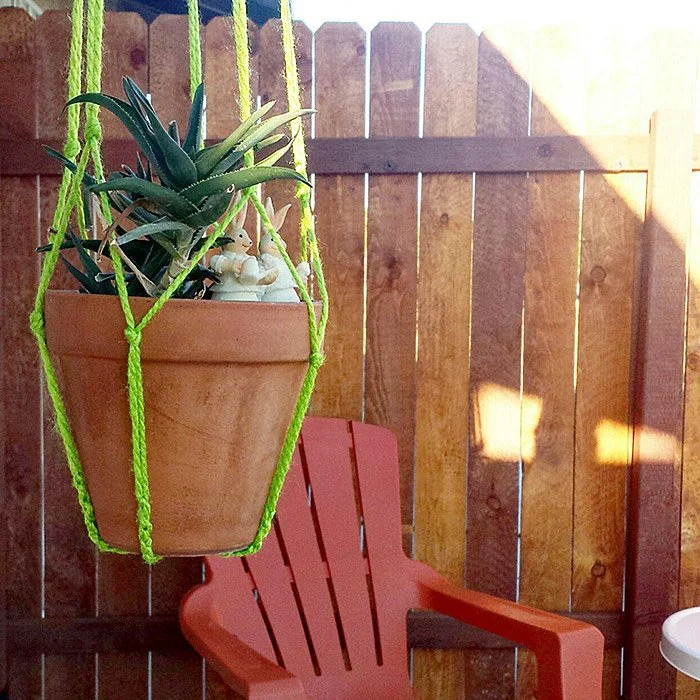 A hanging terracotta pot with a green string holder containing a succulent plant, hanging in front of a wooden fence with sunlight casting shadows. There is an orange plastic chair below the pot.