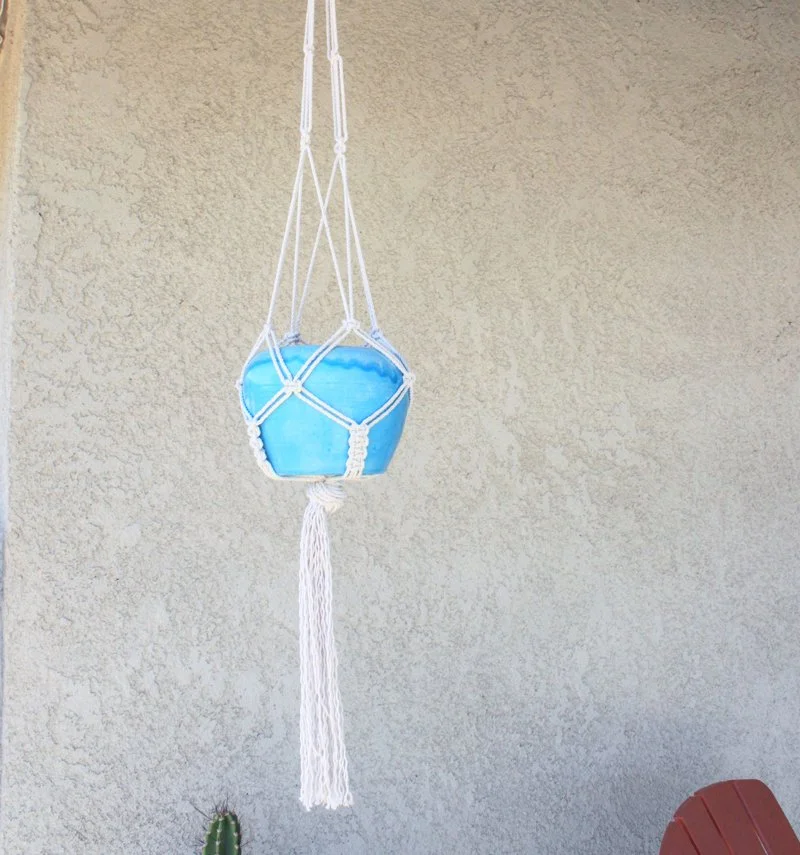 A blue ceramic pot hanging in a white macrame plant holder against a beige textured wall.