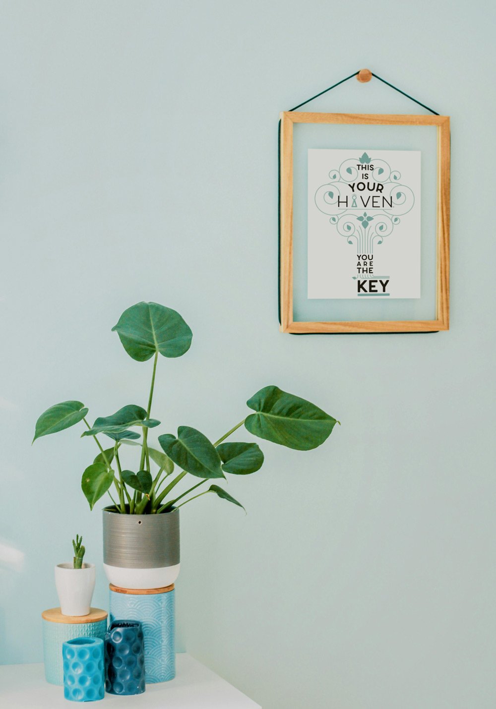 A light green wall with a framed print that says "This is your haven, you are the key" and features decorative swirls and leaves. In front, there are three planters with green plants, including a large plant in a tall, mixed-color pot and two smaller plants in blue and white pots.
