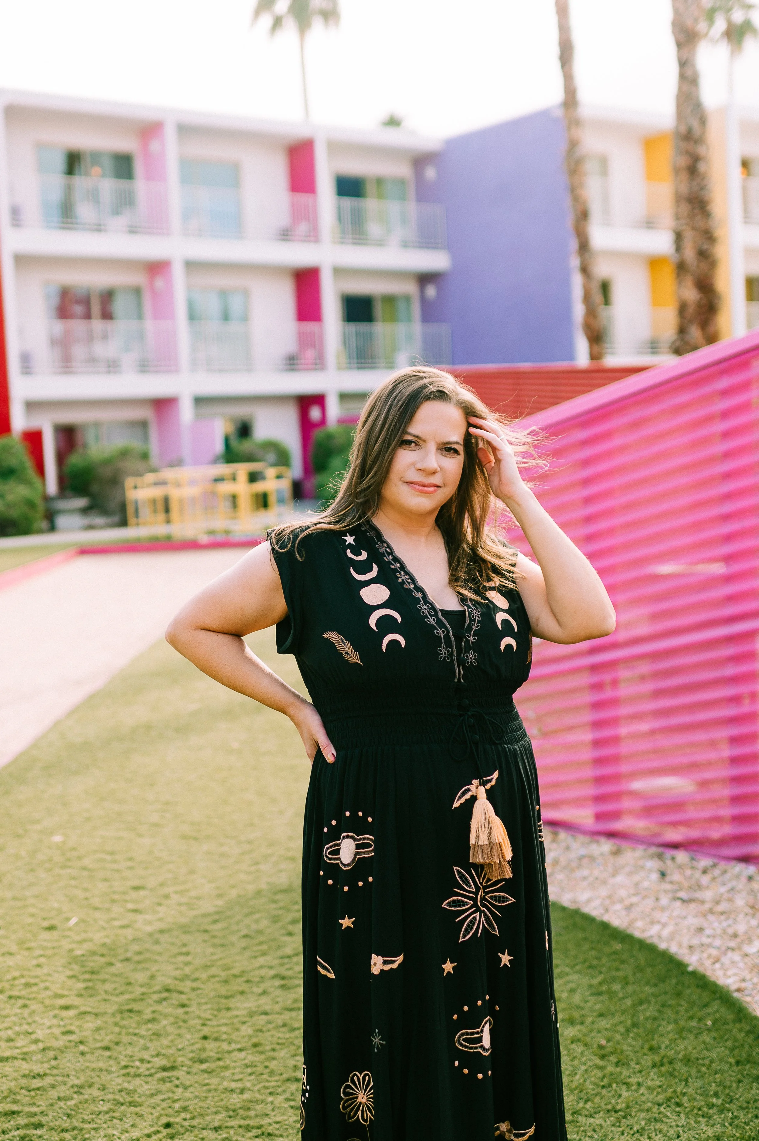 A woman standing outdoors near a pink fence, wearing a black dress with moon and star patterns, with modern colorful apartment buildings and palm trees in the background.