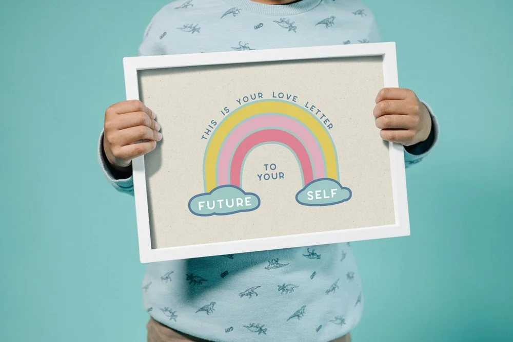 Child holding a framed love letter to their future self featuring a rainbow and the words: "This is your love letter to your future self."