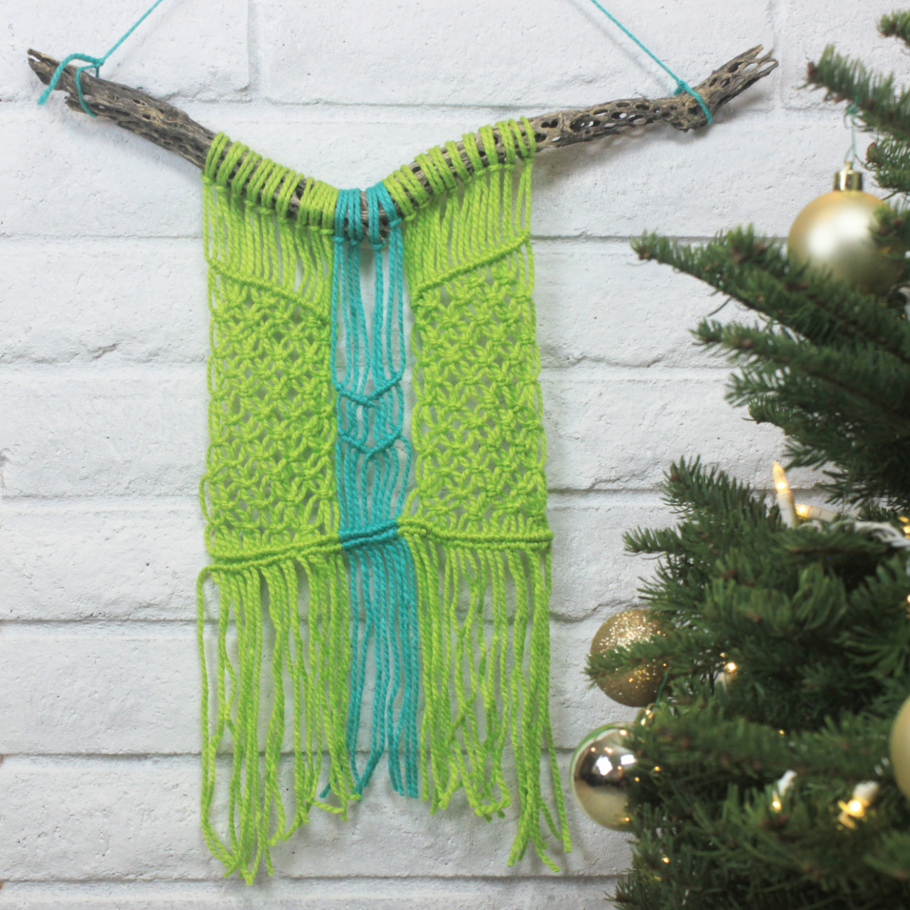 Handmade woven wall hanging with green and blue yarns, draped on a piece of cholla skeleton against a white brick wall, next to decorated Christmas tree with gold ornaments.