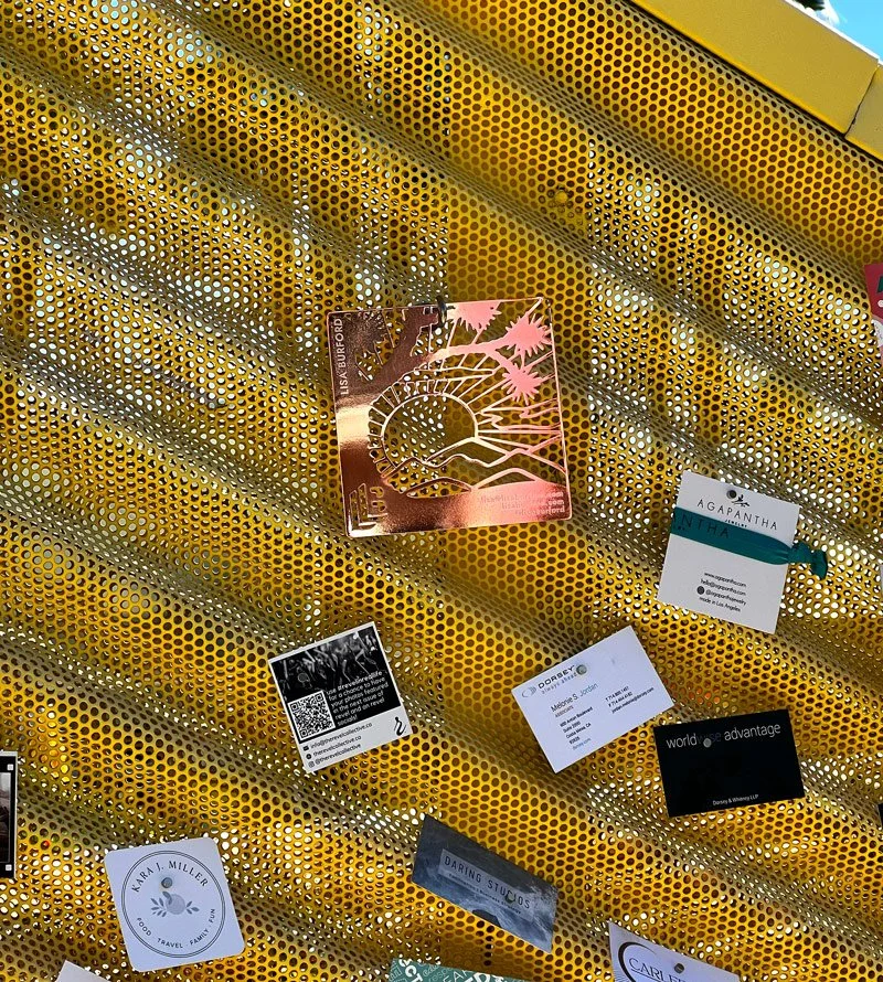 A square metal Lisa Burford business card hangs from a magnetic hook onto a metal mesh-like curvy wall. Other business card designs of varying shapes and sizes are nearby on the wall.