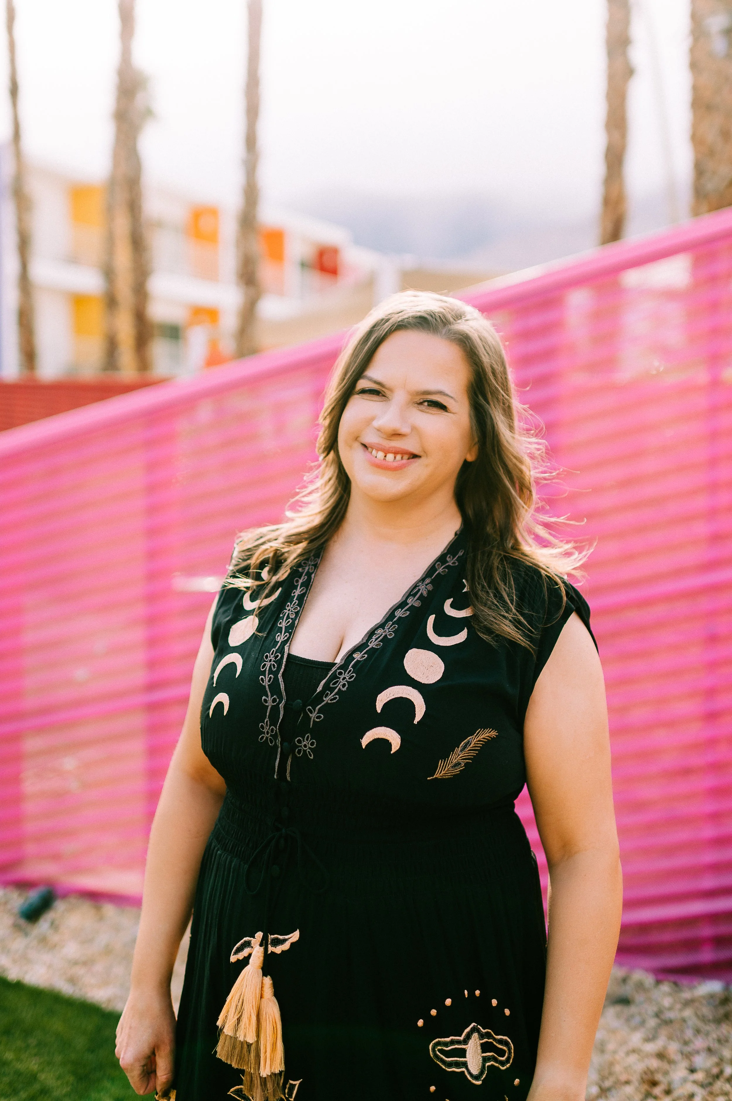 A woman with brown hair wearing a black dress with embroidery, smiling and squinting at the camera, standing outdoors near a pink fence with residential buildings and trees in the background.