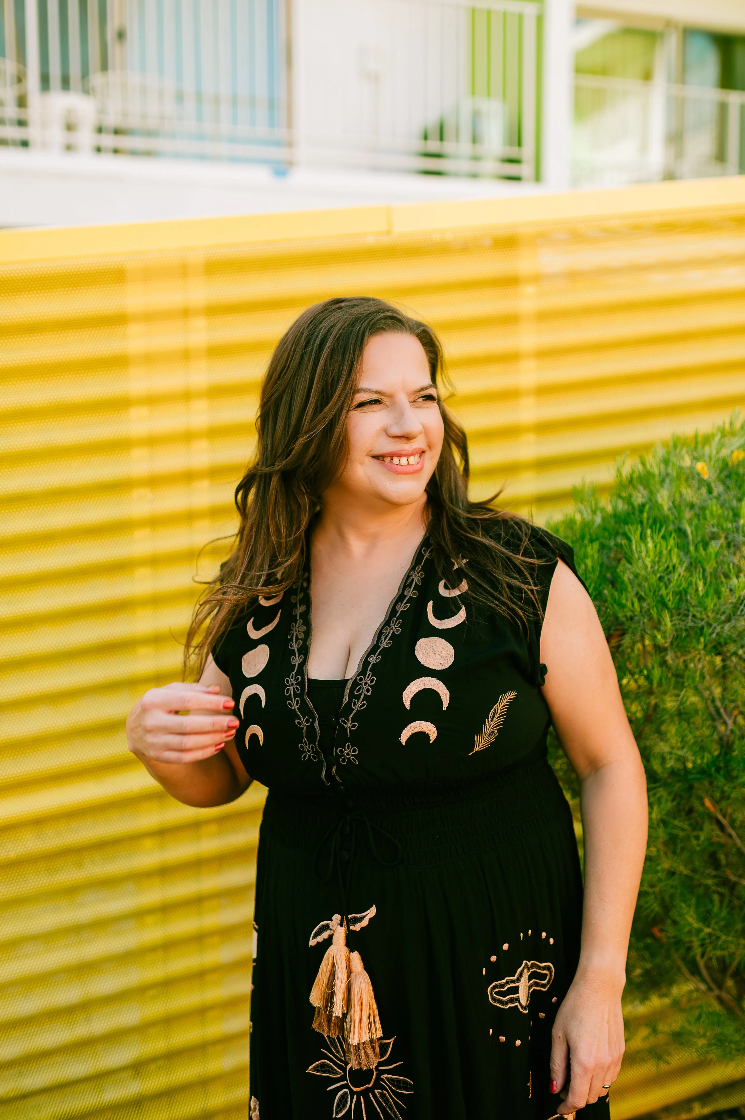 A woman with long brown hair smiling and looking to the side, standing outdoors near a green bush and a yellow fence.