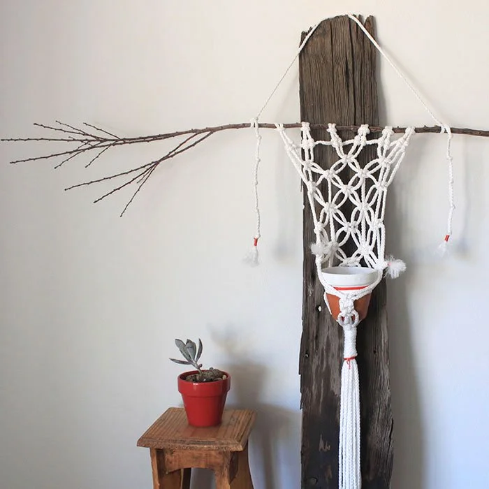 A macrame wall hanging hung from a rustic piece of wood. A small red pot with a plant on a wooden stool sits next to it.