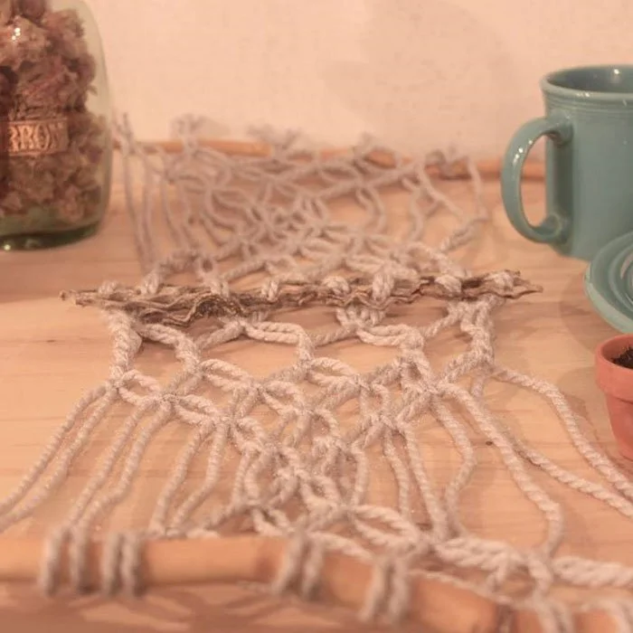 Close-up of a handmade macrame table runner with wooden beads, a stack of books in the background, and a teal coffee mug on a wooden table.