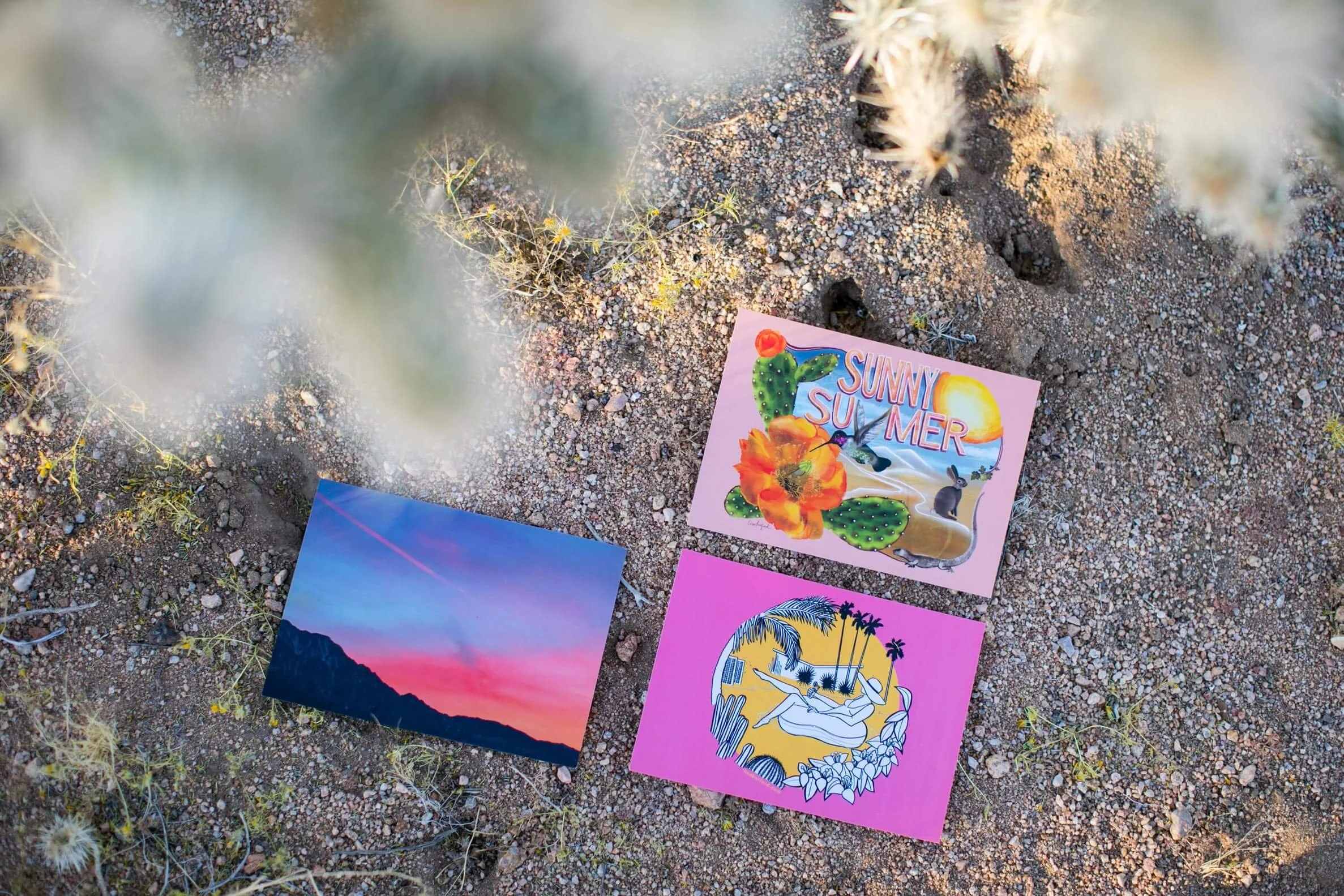 Three colorful postcards on the sand.  1 card shows a sunset over mountains, another has a pink background with a woman and palm trees, and the third has a desert scene with cacti and the words Sunny Summer.