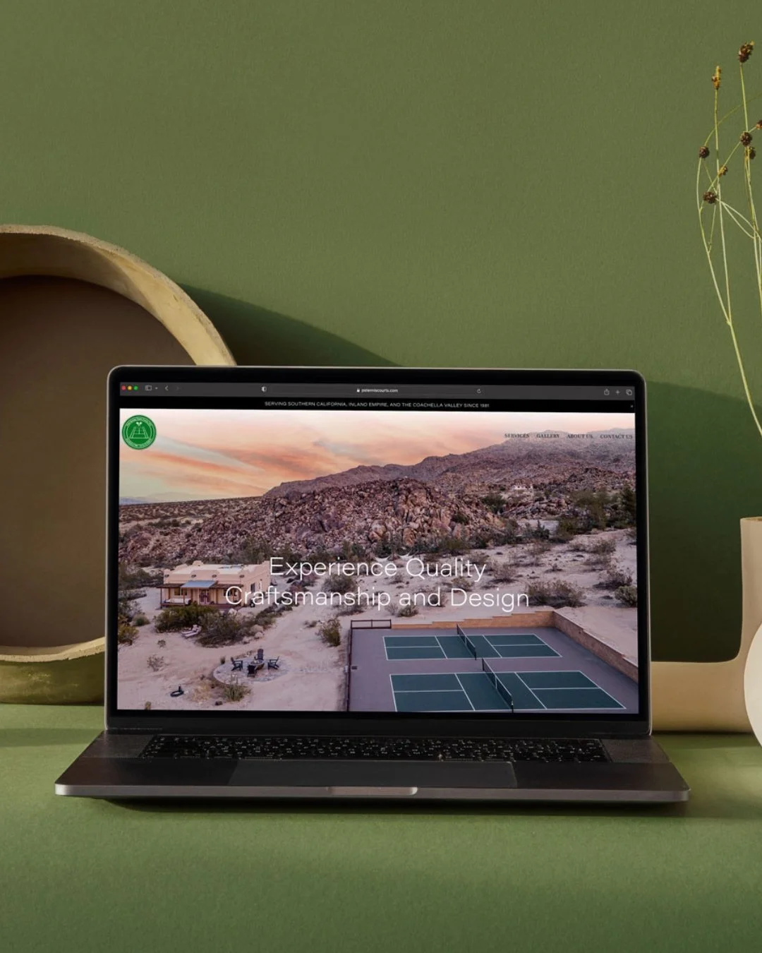 Laptop displaying a desert landscape with mountains and tennis courts on green surface, with a green wall and decorative pottery in the background.