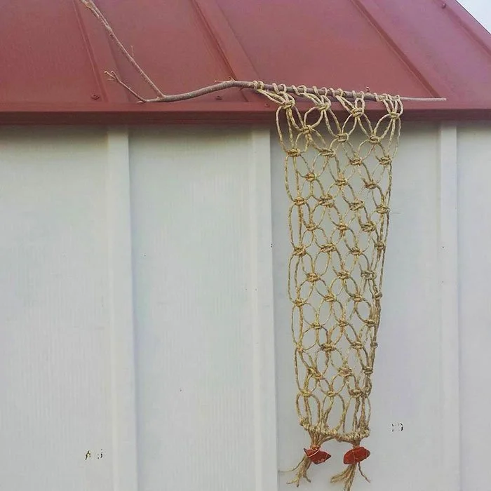 A handmade jute macrame wall hanging hangs from the edge of a shed roof.