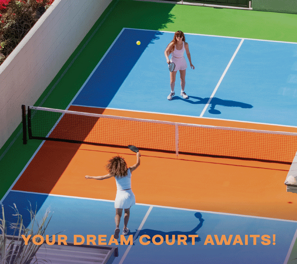 Two women playing tennis on a colorful outdoor court with a net, one preparing to hit the ball and the other standing near the baseline, with text saying 'Your Dream Court Awaits!'