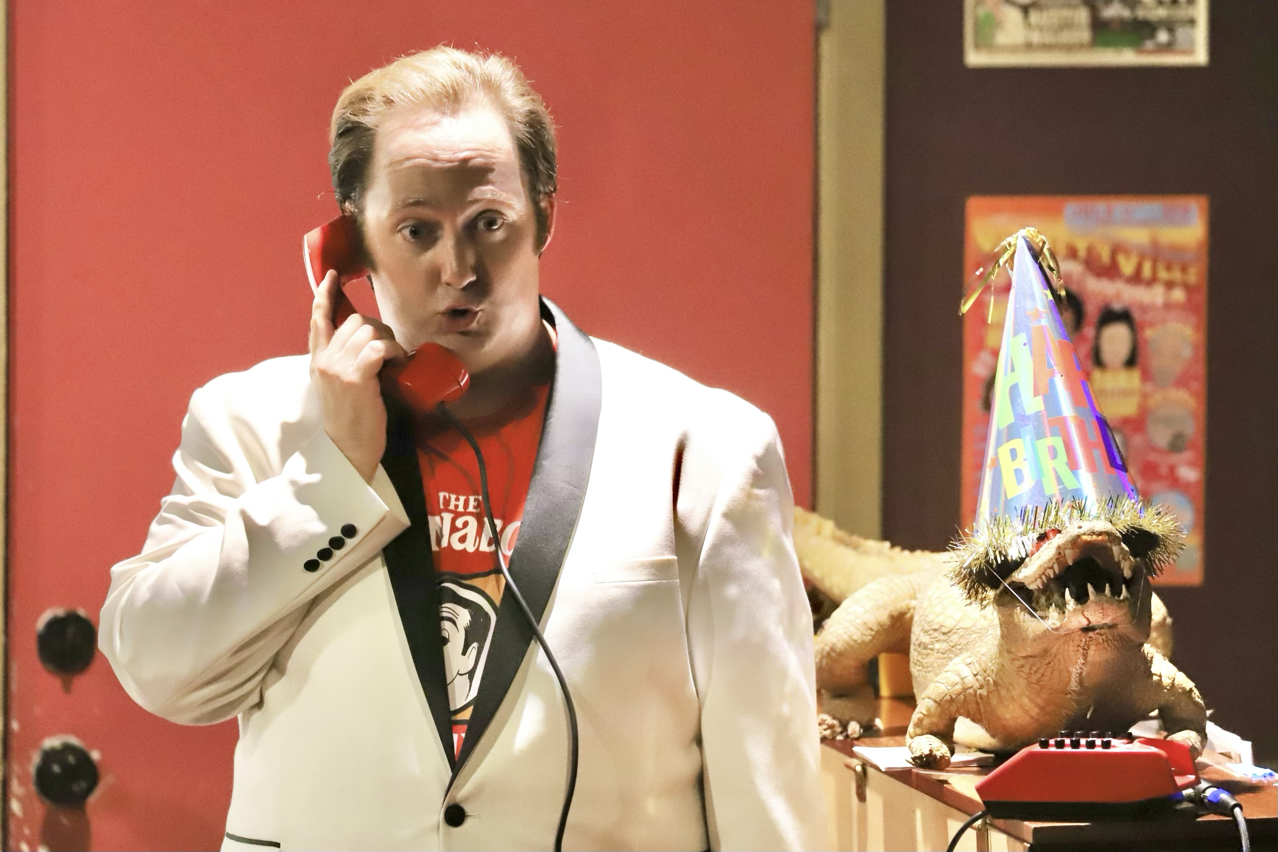 A man in a white tuxedo talking on a red phone, wearing a party hat with an alligator on the table behind him. The alligator is wearing a colorful birthday hat and has an open mouth with visible teeth, in a festive setting.