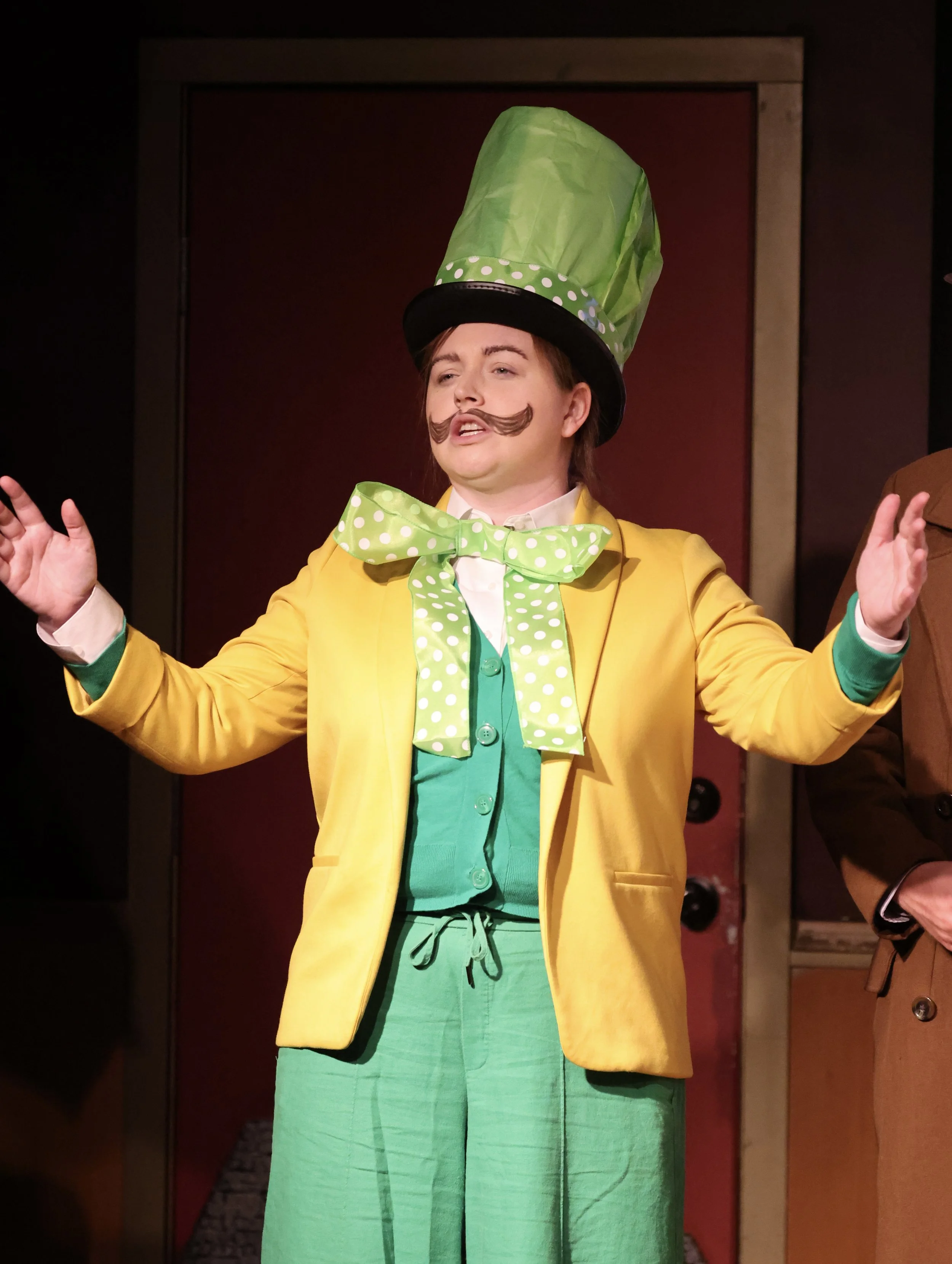 Person in theatrical costume with yellow jacket, green tall hat and pants, and a polka-dot bow tie. Thick mustache drawn on face, performing on stage.
