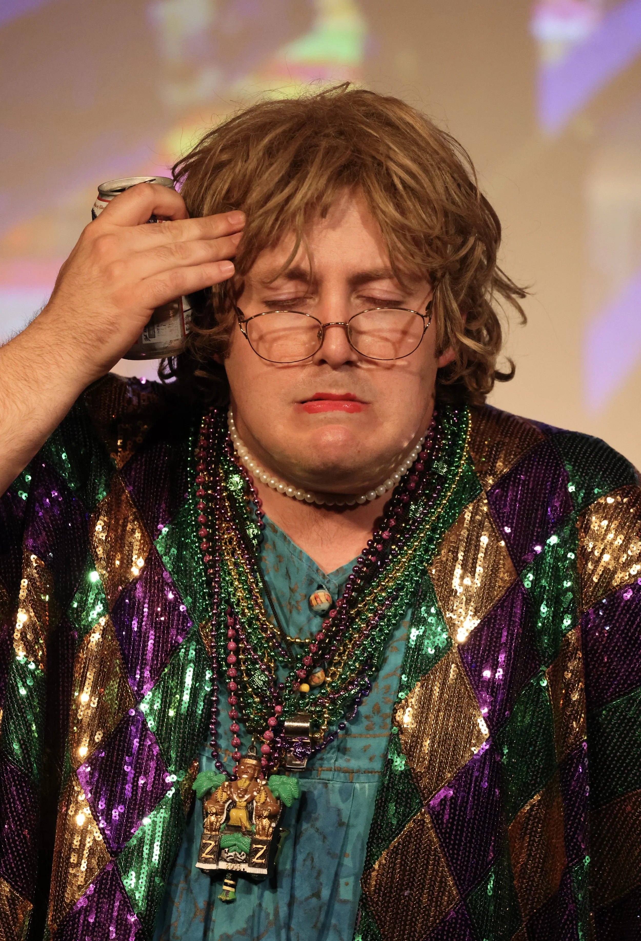 A person with curly hair and glasses, dressed in colorful, shiny clothing and multiple beaded necklaces, holding a can to their head with eyes closed.