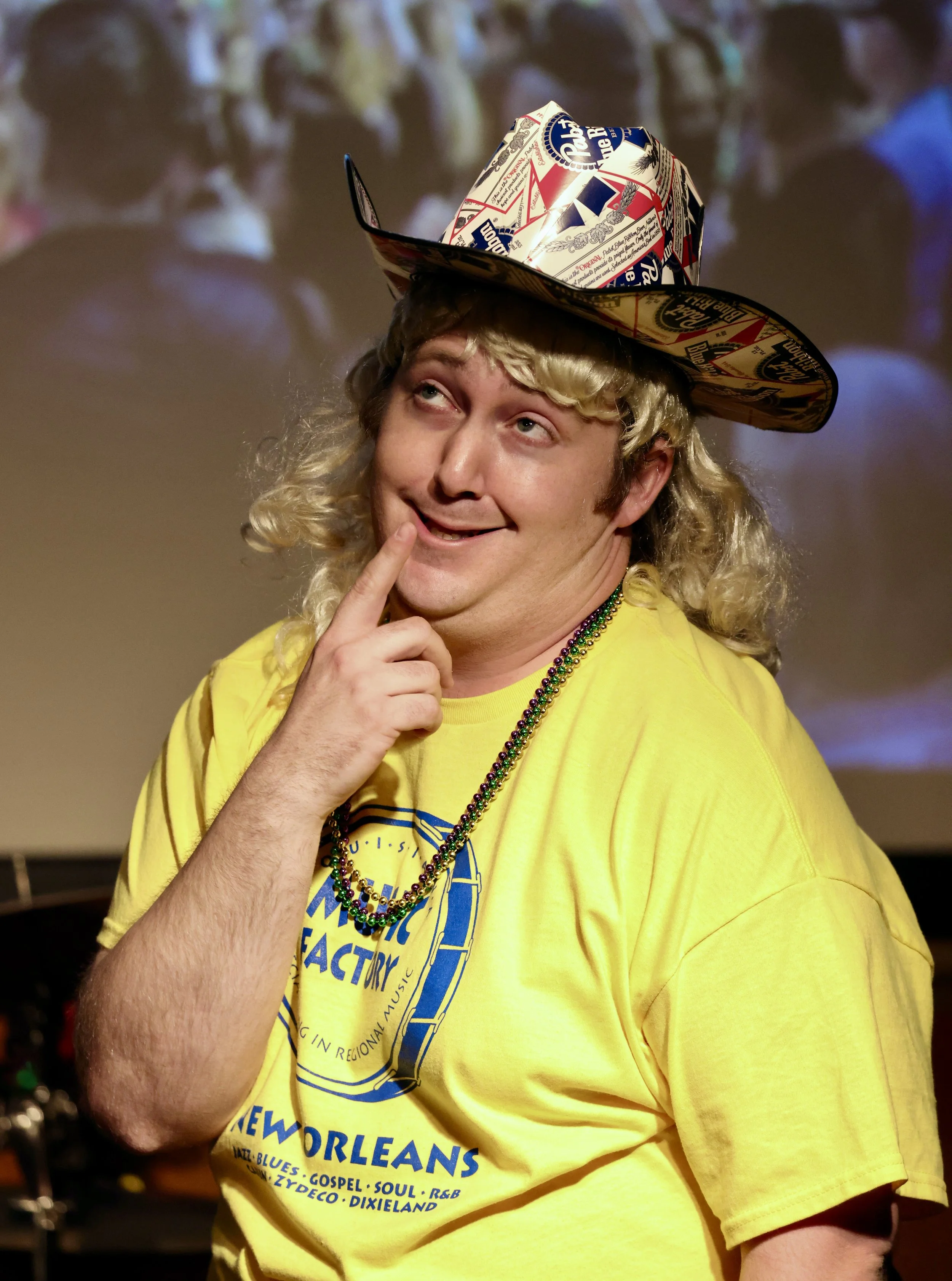 Person with long curly blonde hair wearing a yellow T-shirt with New Orleans design, Mardi Gras beads, and a hat made from Pabst Blue Ribbon beer packaging, posing with a playful expression.