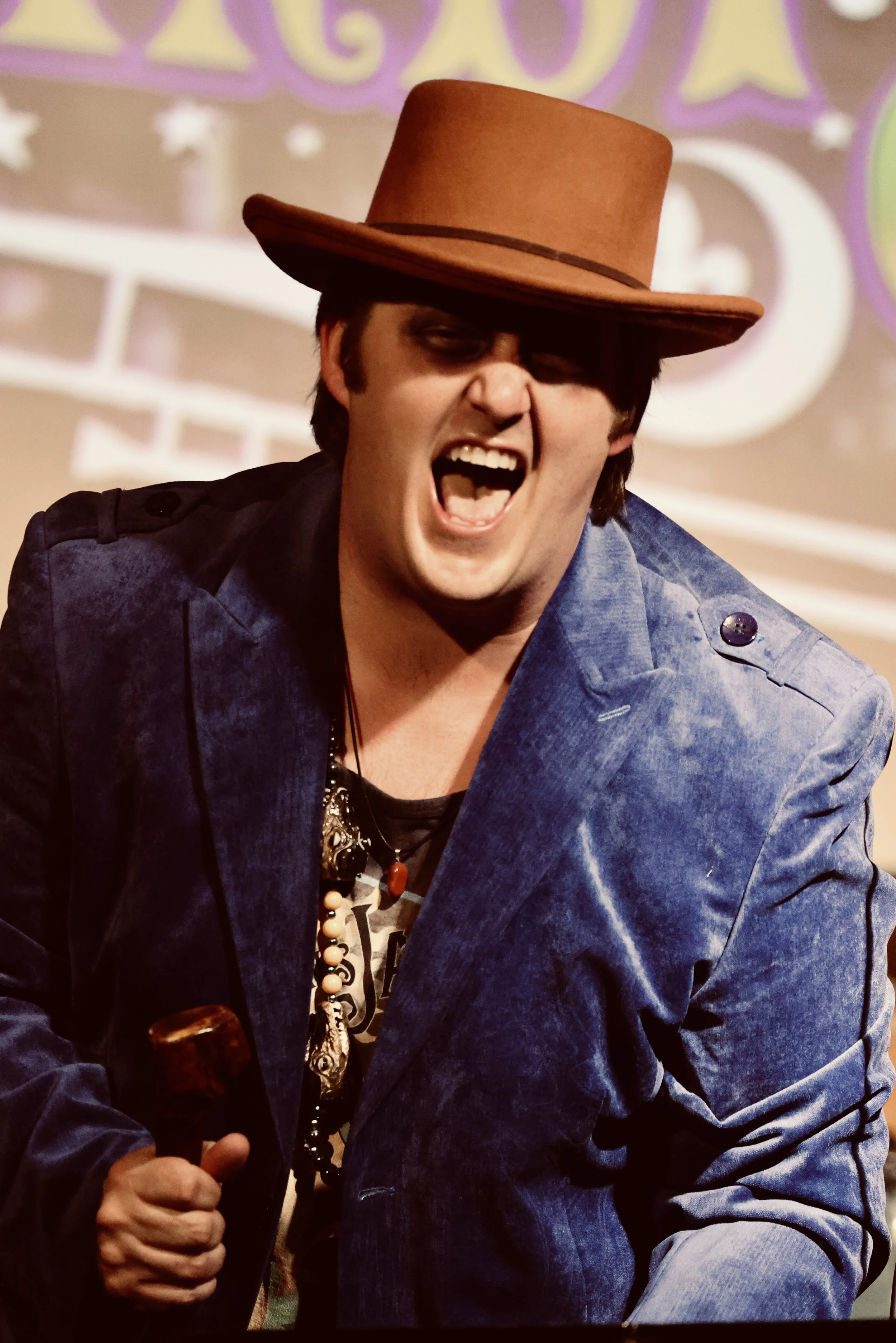 A man wearing a brown hat, blue velvet jacket, and necklaces, appears to be singing or shouting passionately, holding a pipe in his hand.
