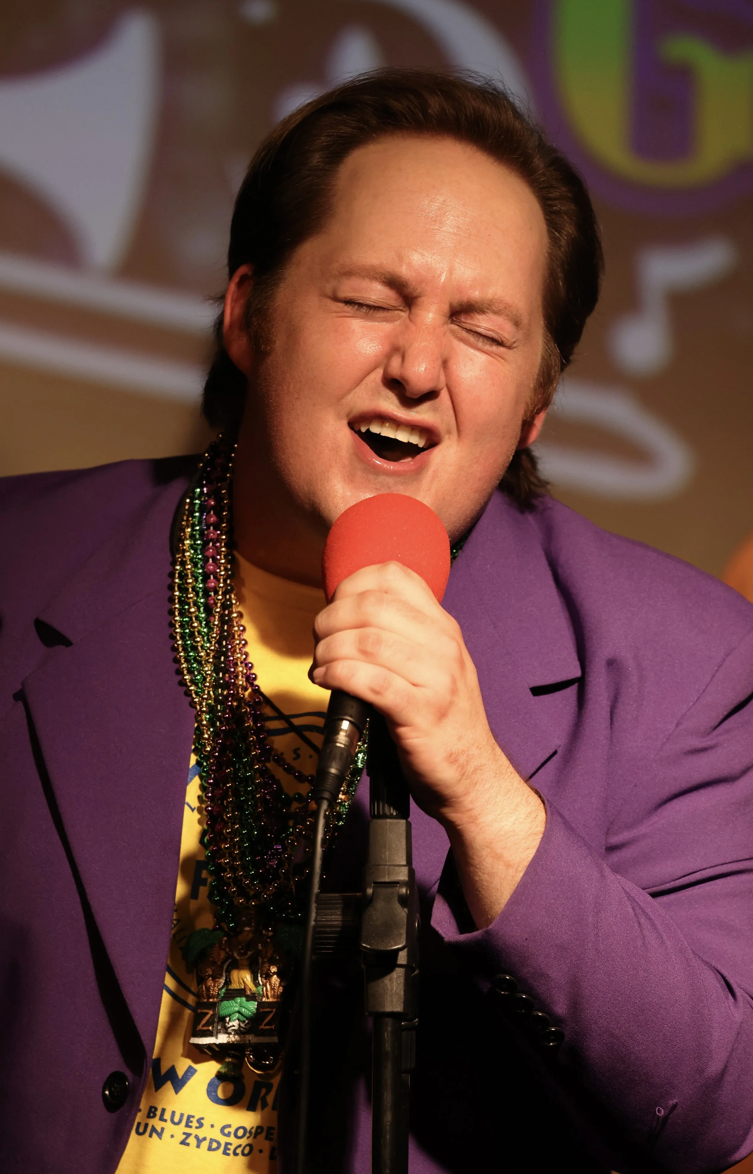 A man singing into a red microphone with eyes closed, wearing a purple jacket, multiple beads, and a yellow shirt, with a colorful background.