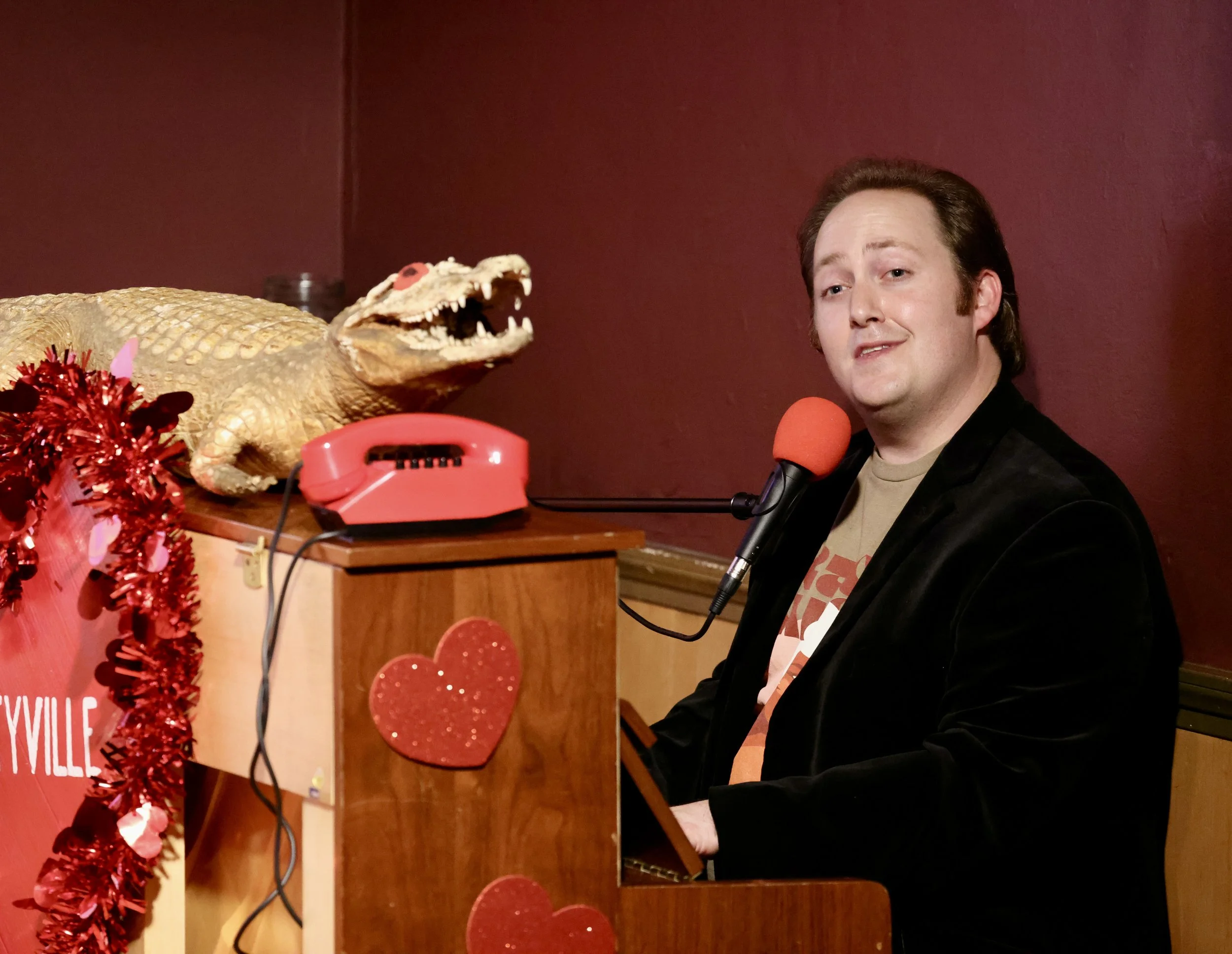 A man with brown hair wearing a beige T-shirt and black blazer, sitting at a piano with a red microphone, smiling slightly, with a dragon head prop and some Valentine's Day decorations on the piano, against a red background.