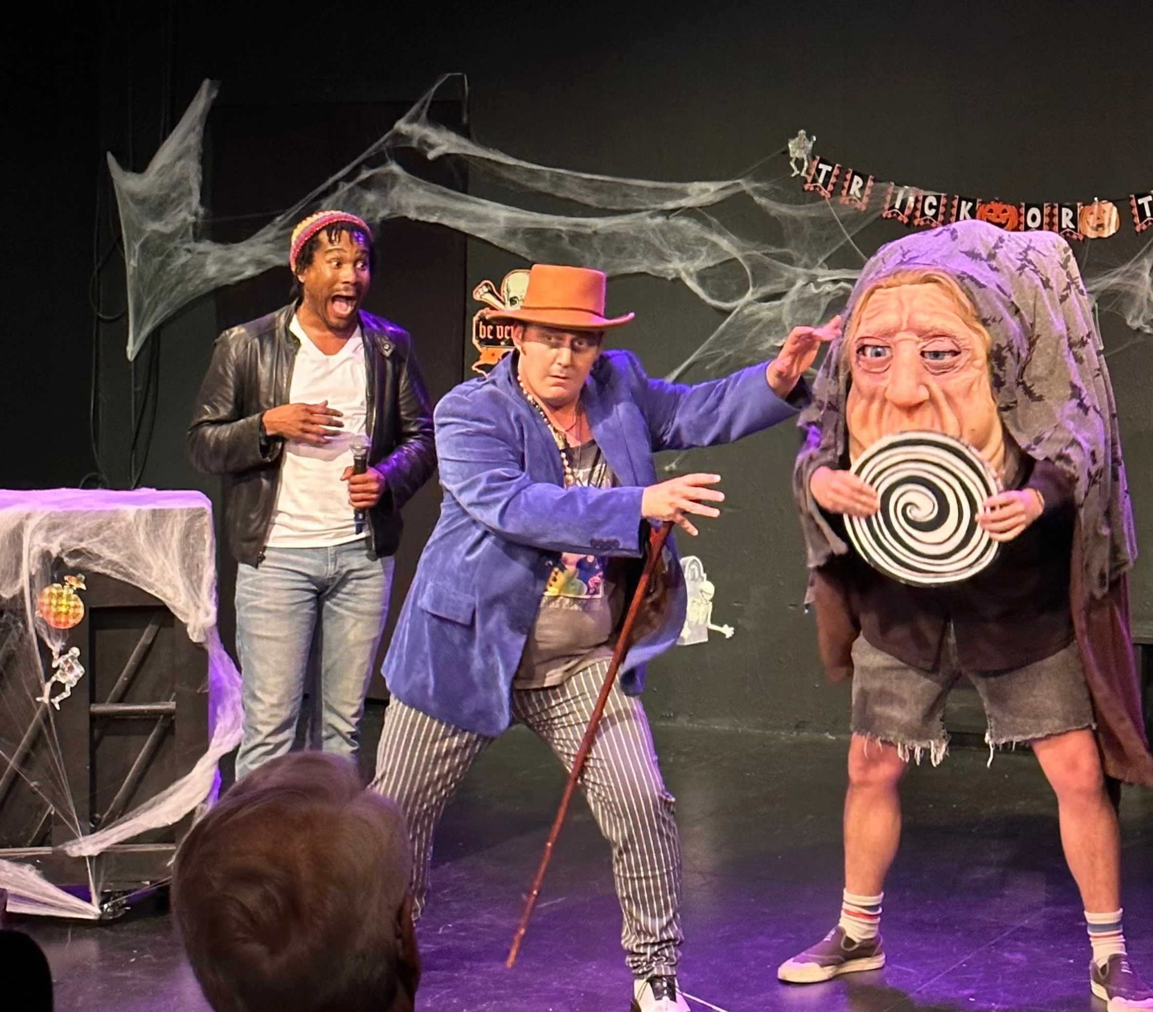 Three performers dressed in Halloween costumes on a stage with spider webs and a Halloween banner in the background. One performer wears a leather jacket and has dreadlocks, another is dressed as a wizard or magician with a purple coat, striped pants