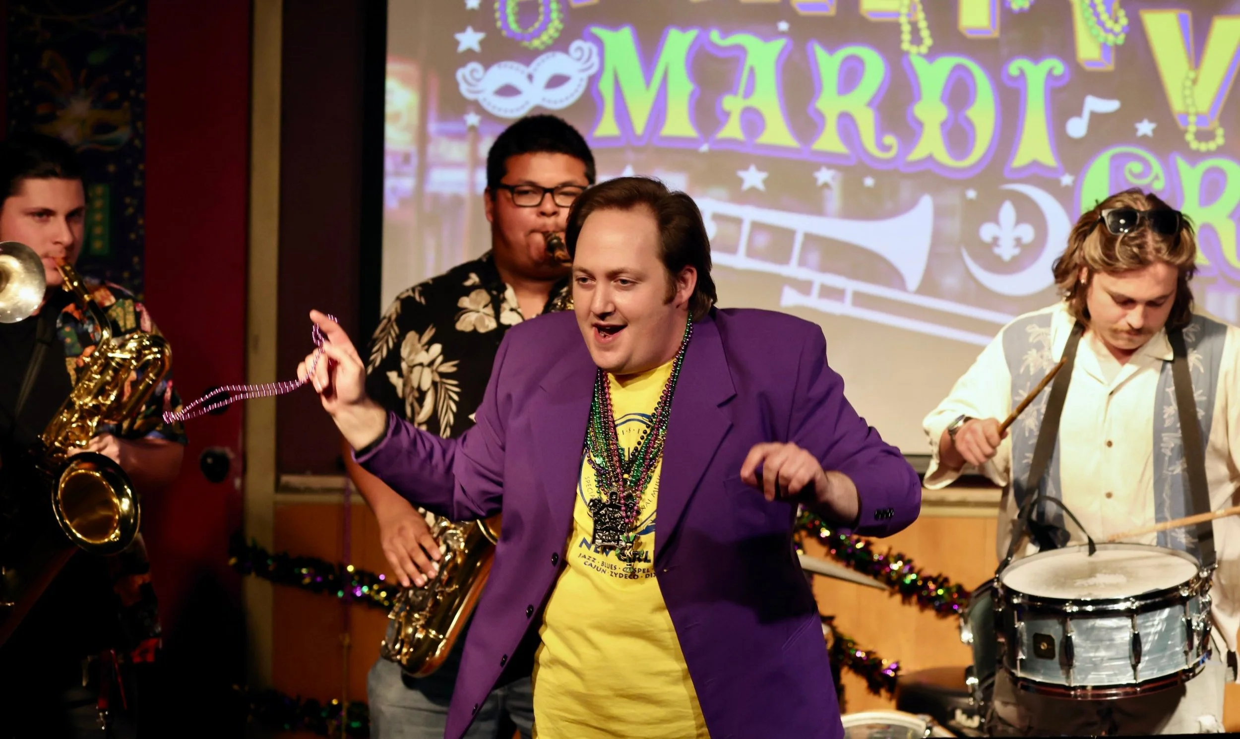 Group of musicians performing at a Mardi Gras event, one wearing a purple blazer and beads.