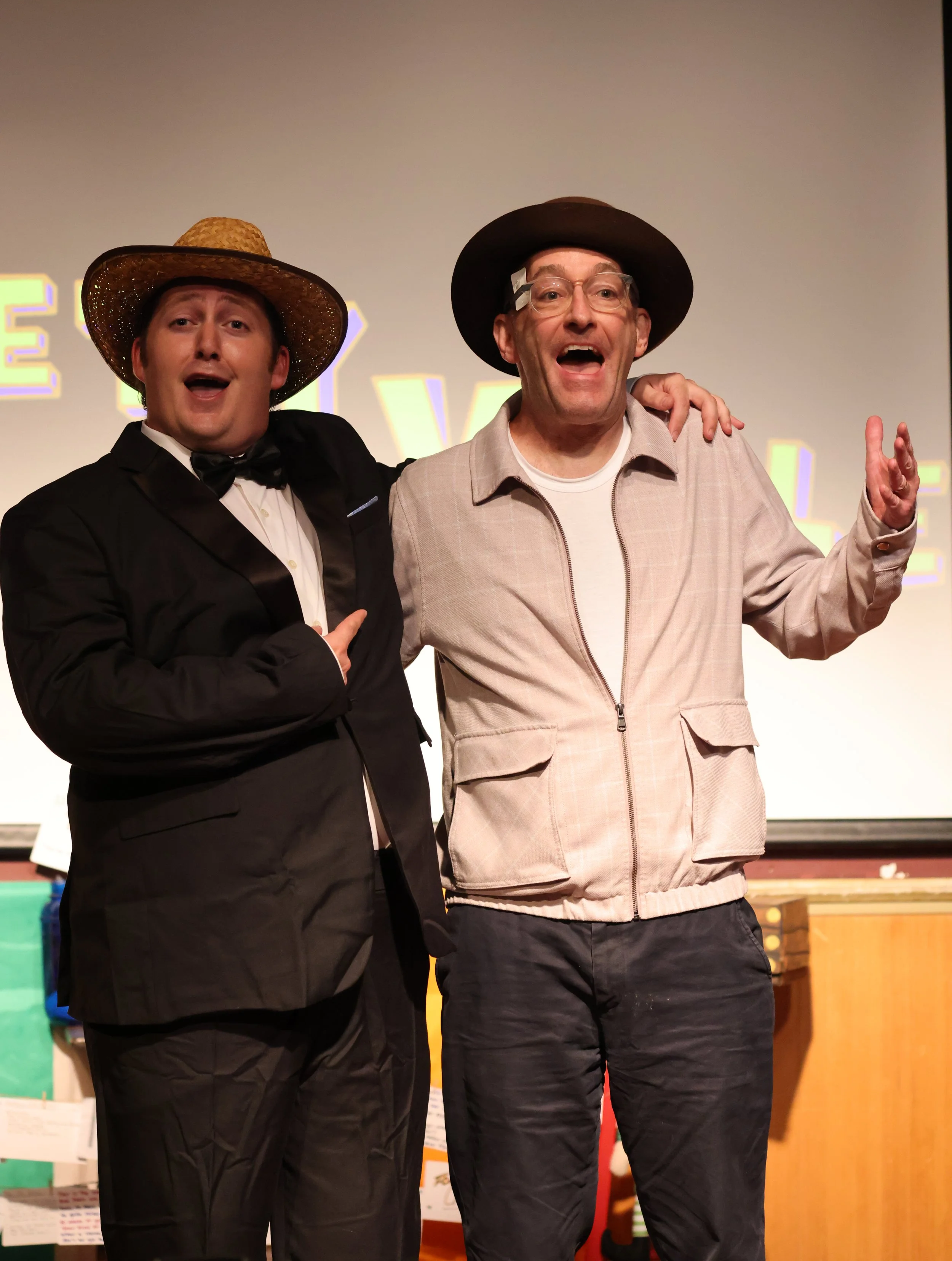 Two men in costumes performing on stage, one in a tuxedo with a large straw hat, the other in a beige jacket and a dark fedora, smiling and gesturing.