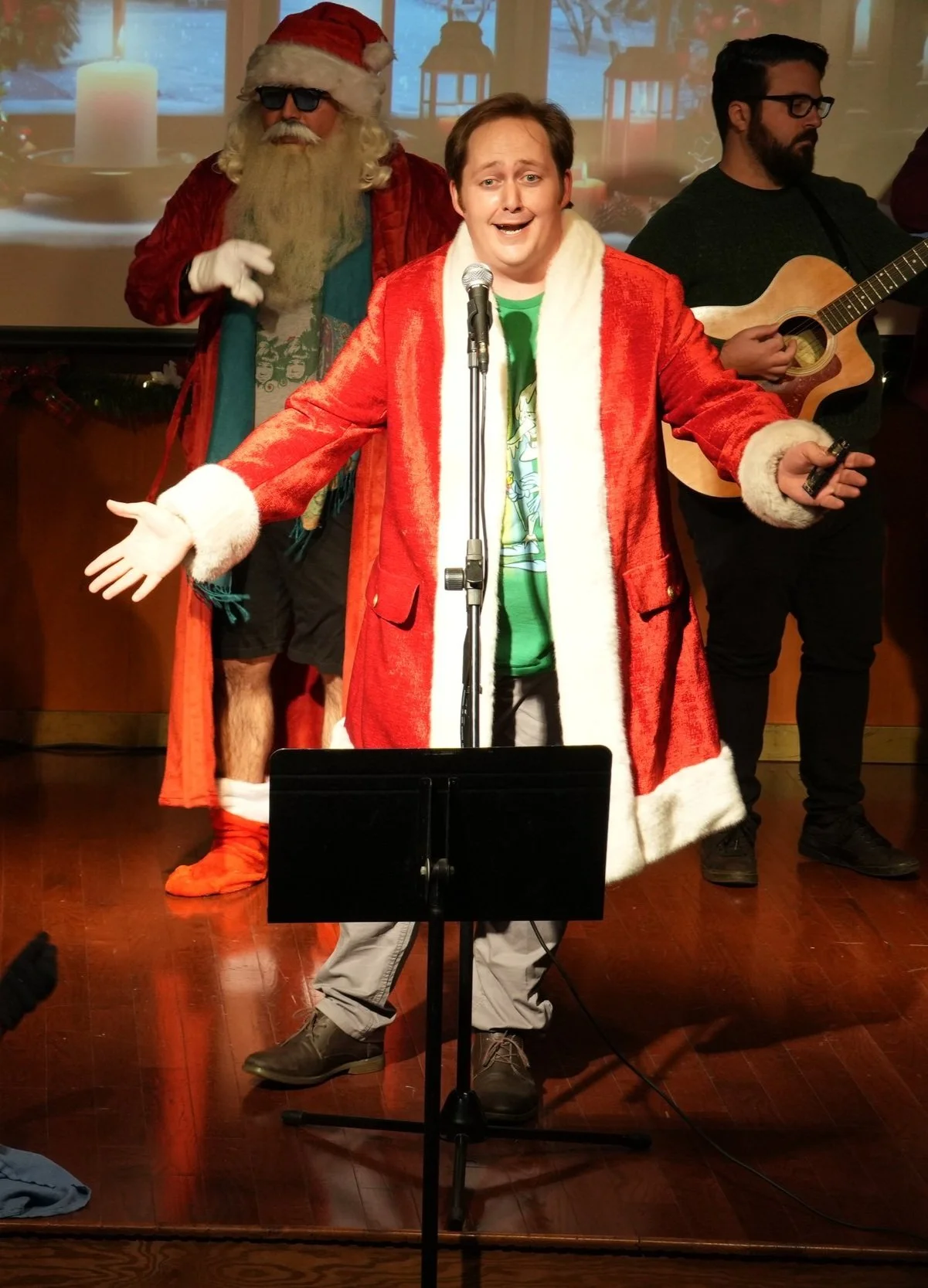 Man dressed as Santa Claus with a red coat, standing in front of a microphone, singing on stage during a Christmas event, with two musicians behind him; one with sunglasses and a beard in a Santa hat and red coat, the other playing a guitar and weari