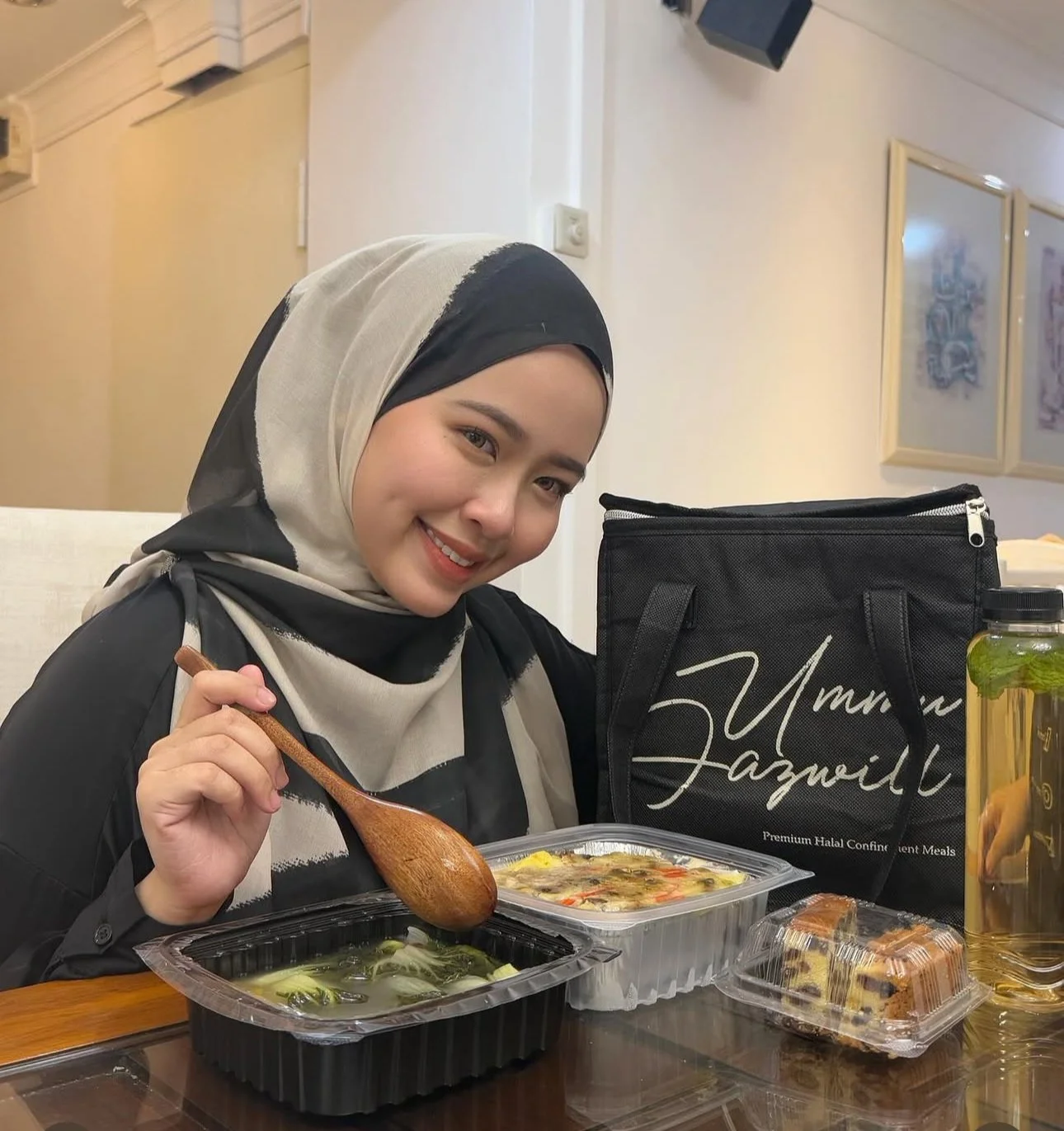 Sonia Asyira wearing a black and white hijab sitting at a table with her confinement meals