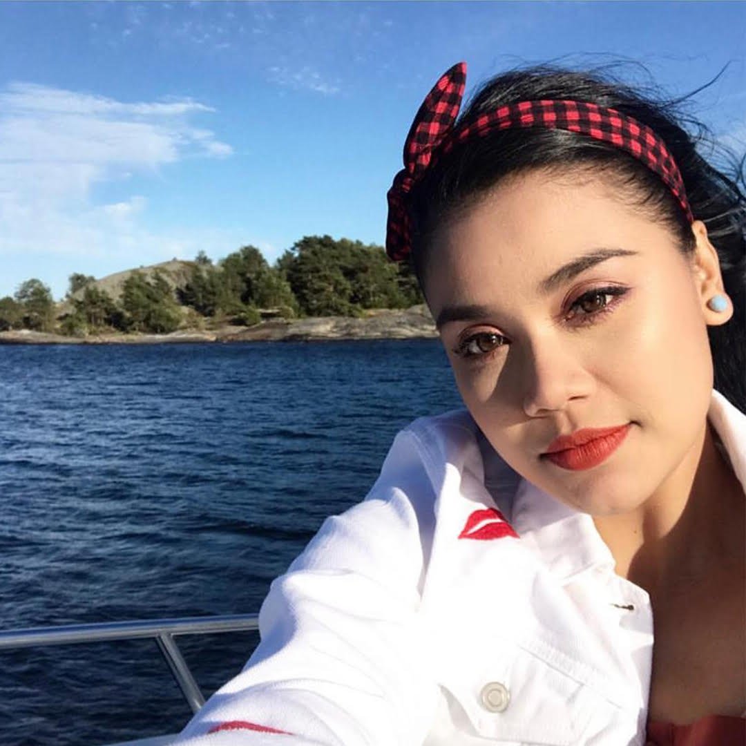 Huda Ali, wearing a red and black checkered headband and a white jacket, taking a selfie on a boat with water and trees in the background.