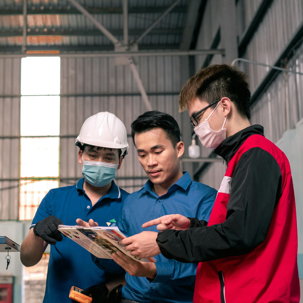 3 men looking at an ipad, 2 wear masks and one wearing a helmet