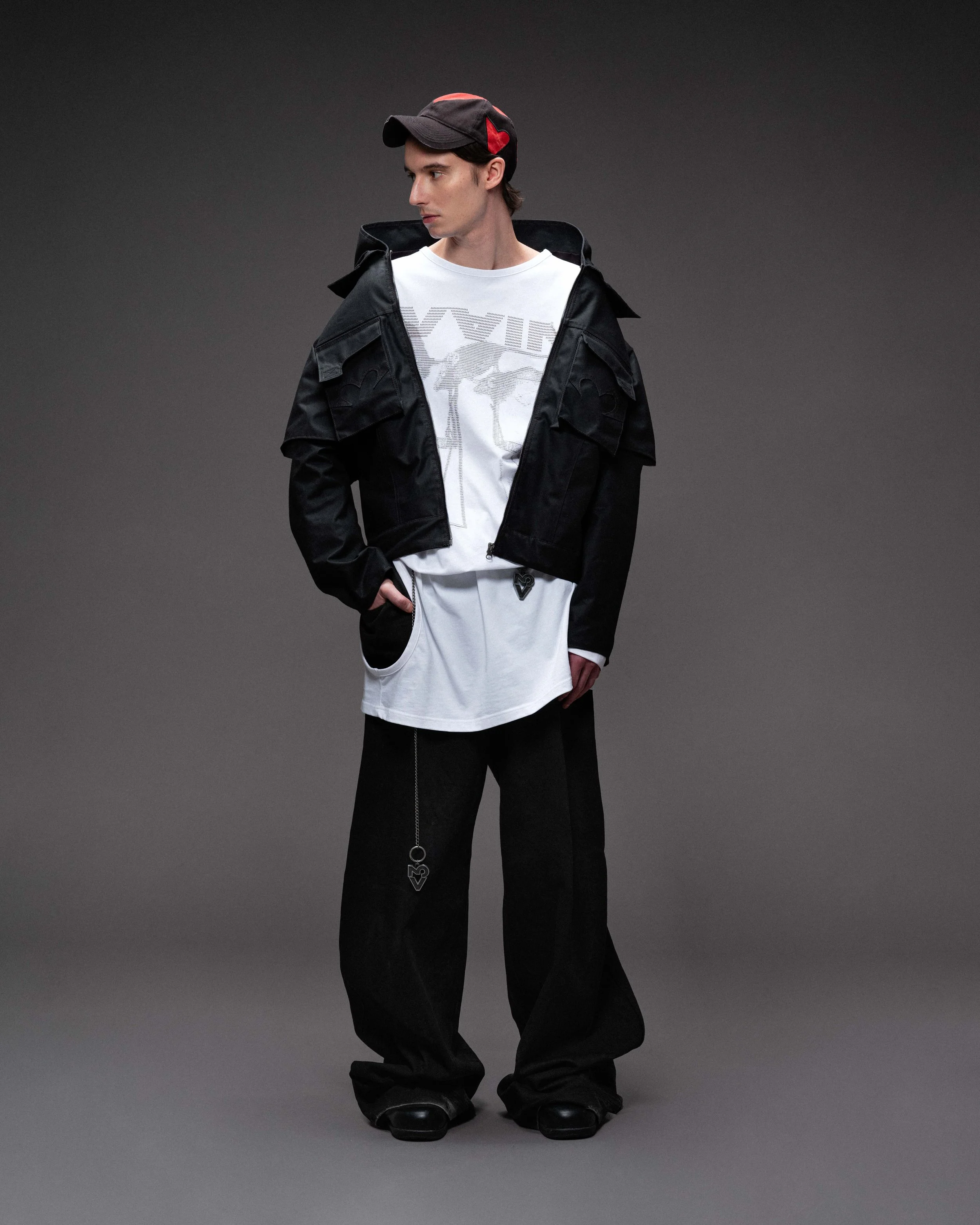 A young man wearing a black cap, black jacket, white graphic t-shirt, black baggy pants, and black shoes standing against a gray background. VAIN