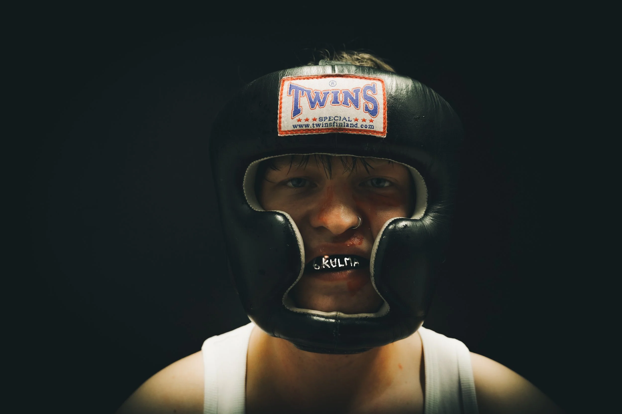 A young male boxer wearing a black protective headgear with "Twins" brand label, looking directly at the camera against a dark background.